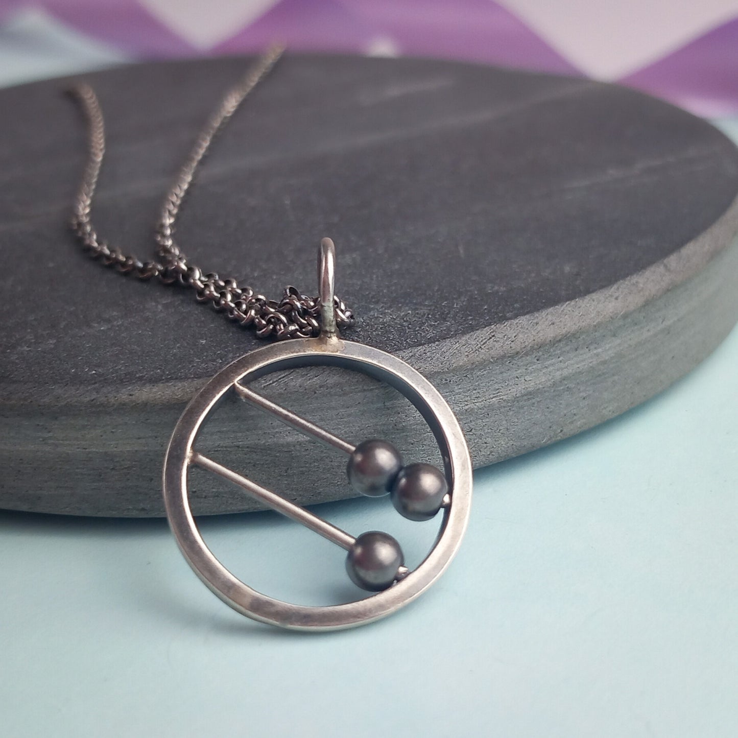 Sterling silver abacus-style fidget pendant necklace by Rebecca Davies Jewellery. The circular pendant features two parallel bars with movable dark metal beads, creating an interactive, minimalist design. Displayed on a dark slate surface with a delicate silver chain, the pendant combines modern aesthetics with tactile functionality. Ideal for fidgeting or meditation jewellery lovers.