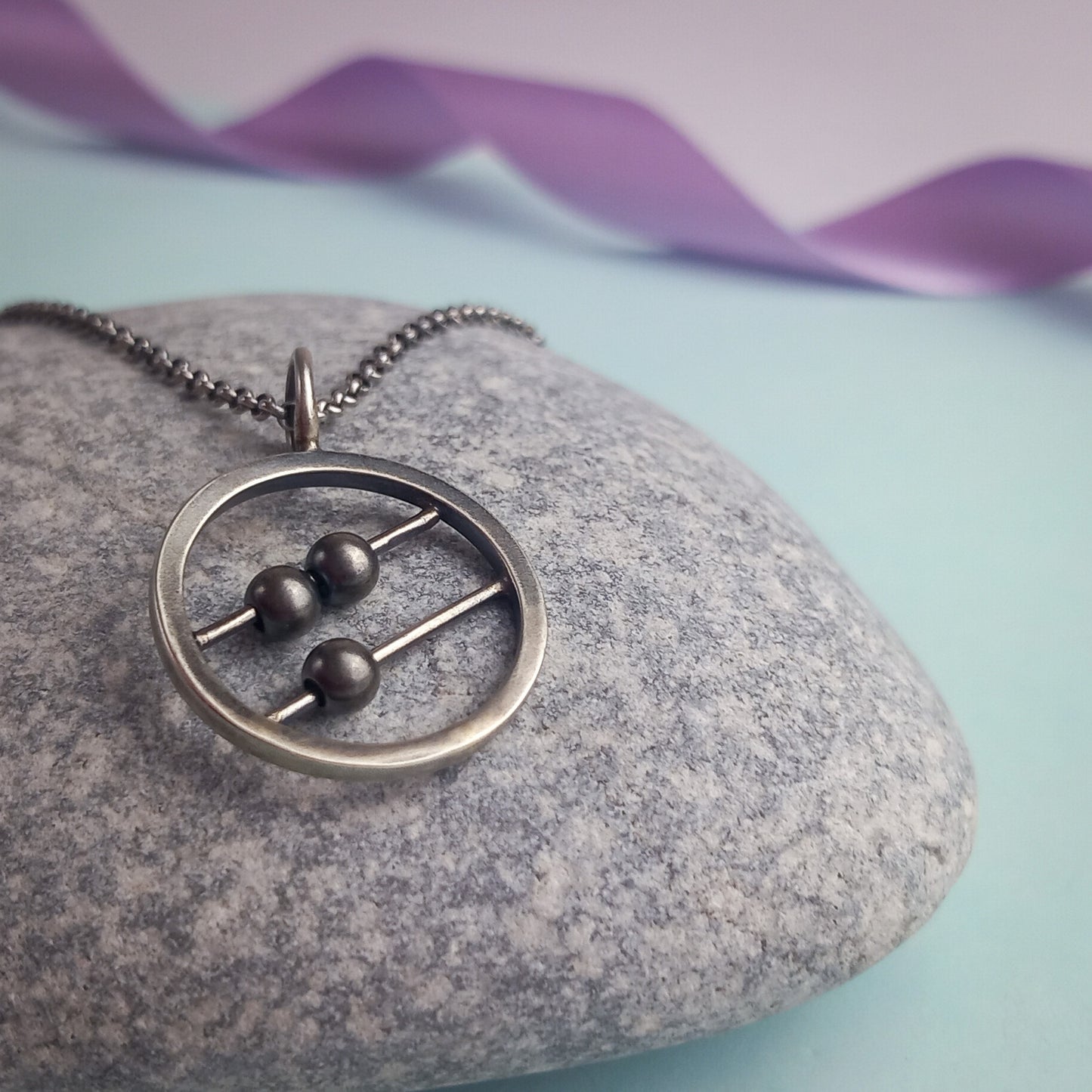 Sterling silver abacus-style fidget pendant necklace by Rebecca Davies Jewellery. The circular pendant features two parallel bars with movable dark metal beads, creating an interactive, minimalist design. Displayed on a grey stone surface with a delicate silver chain, the pendant combines modern aesthetics with tactile functionality. Ideal for fidgeting or meditation jewellery lovers.