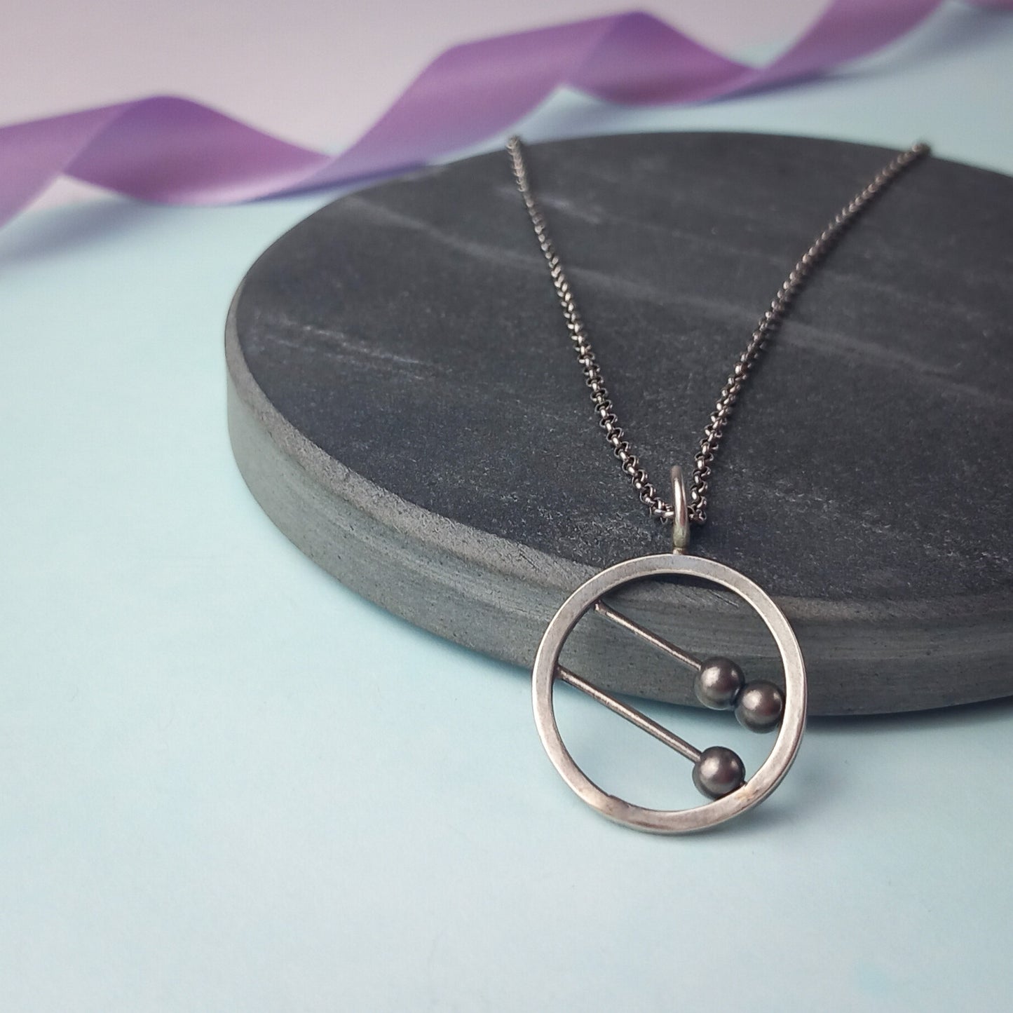 Sterling silver abacus-style fidget pendant necklace by Rebecca Davies Jewellery. The circular pendant features two parallel bars with movable dark metal beads, creating an interactive, minimalist design. Displayed on a dark slate surface with a delicate silver chain, the pendant combines modern aesthetics with tactile functionality. Ideal for fidgeting or meditation jewellery lovers.