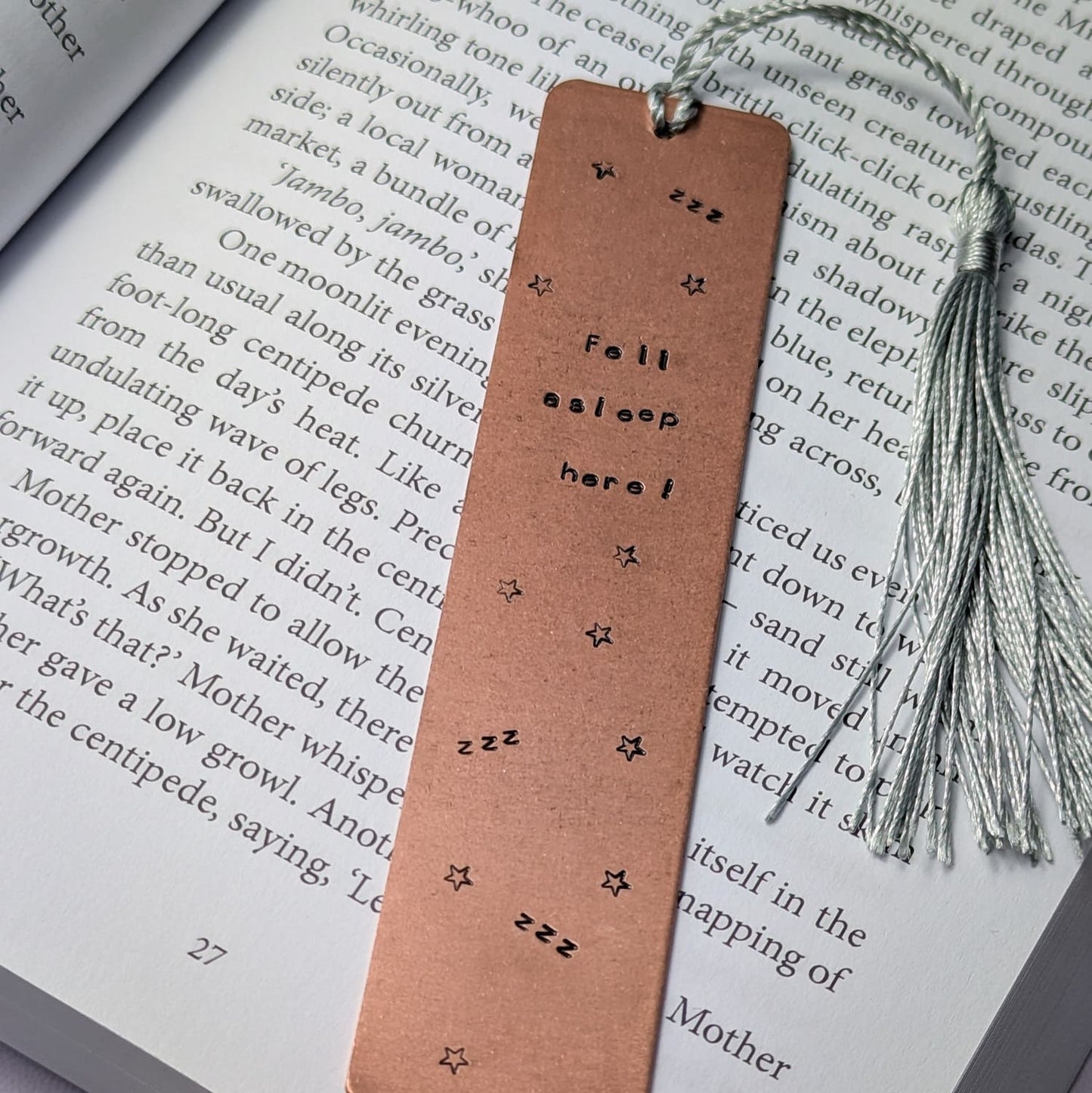 Handmade copper bookmark with "Fell Asleep Here" hand-stamped in black lettering, featuring little Z's and stars stamped throughout. A light grey tassel is attached, resting on an open book with printed text. Perfect literary gift for book lovers and avid readers