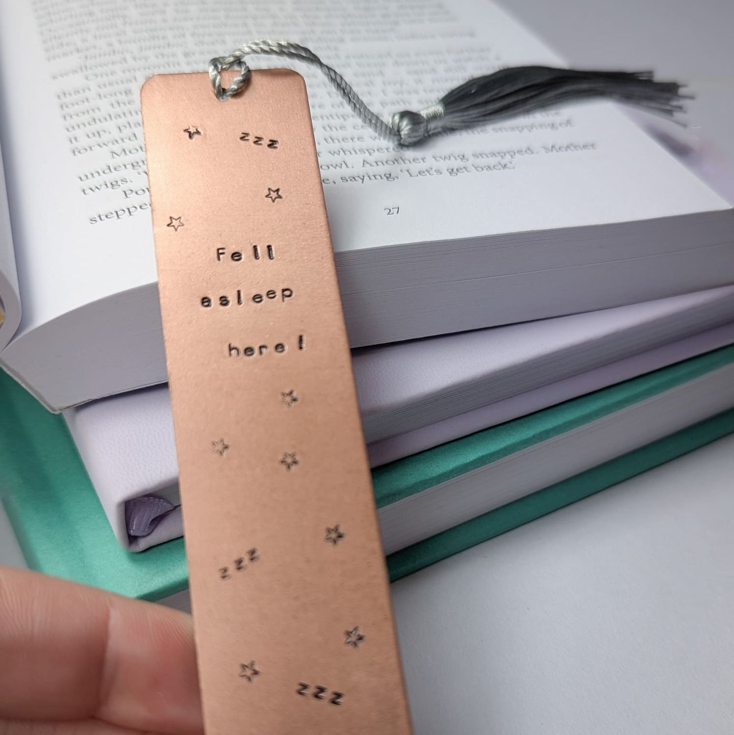 Handmade copper bookmark with "Fell Asleep Here" hand-stamped in black lettering, featuring little Z's and stars stamped throughout. A light grey tassel is attached, resting on an open book with printed text. Perfect literary gift for book lovers and avid readers