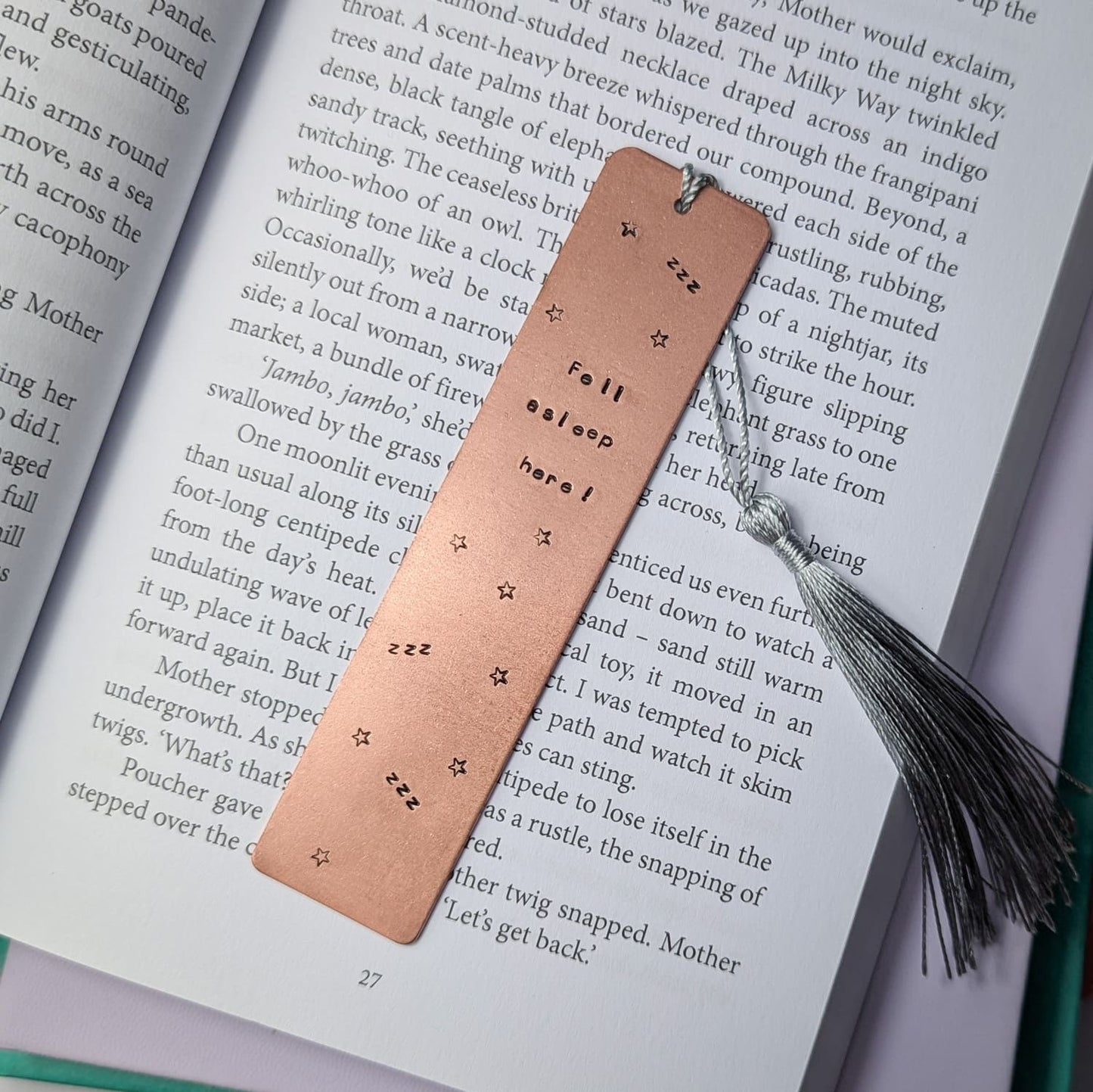 Handmade copper bookmark with "Fell Asleep Here" hand-stamped in black lettering, featuring little Z's and stars stamped throughout. A light grey tassel is attached, resting on an open book with printed text. Perfect literary gift for book lovers and avid readers
