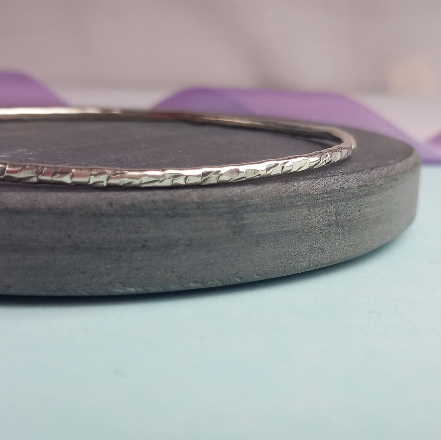Image shows a heavily textured silver bangle sitting on a piece of dark grey slate. In the background is a lavender ribbon, adding a touch of colour.