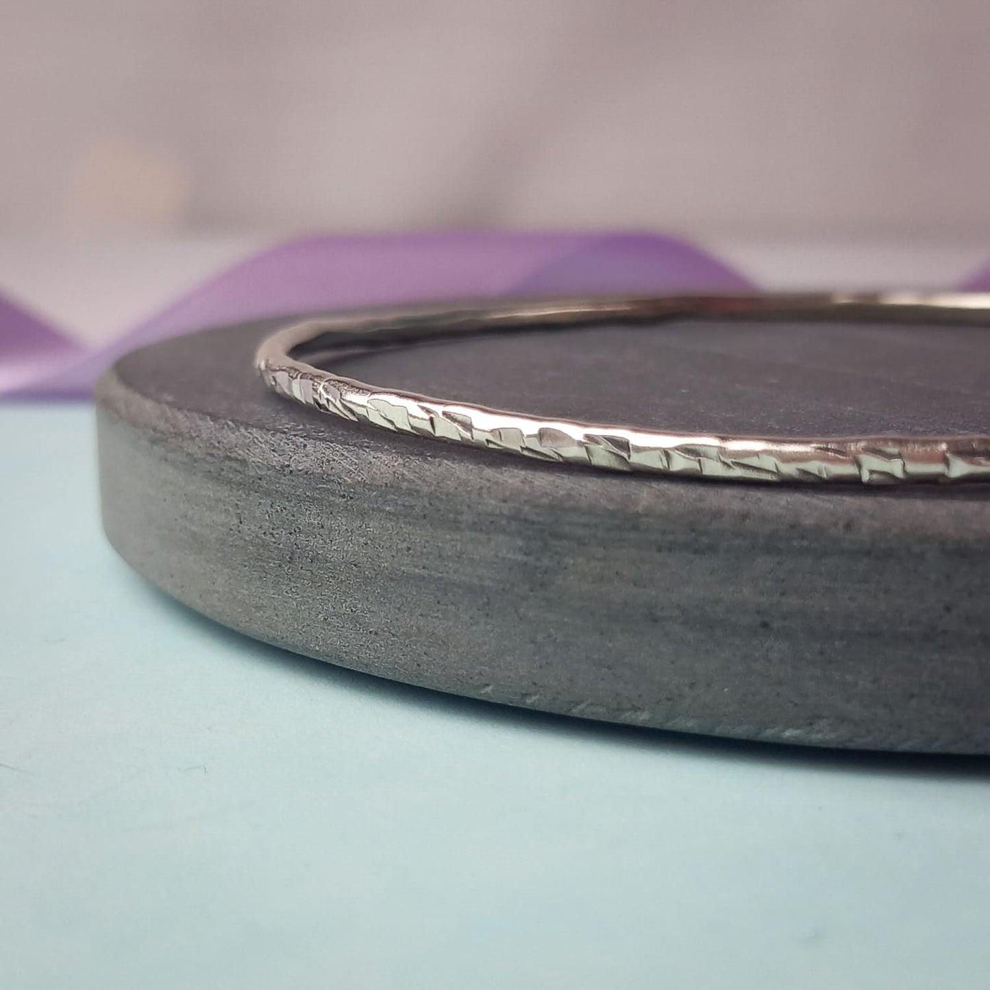 Image shows a heavily textured silver bangle sitting on a piece of dark grey slate. In the background is a lavender ribbon, adding a touch of colour.