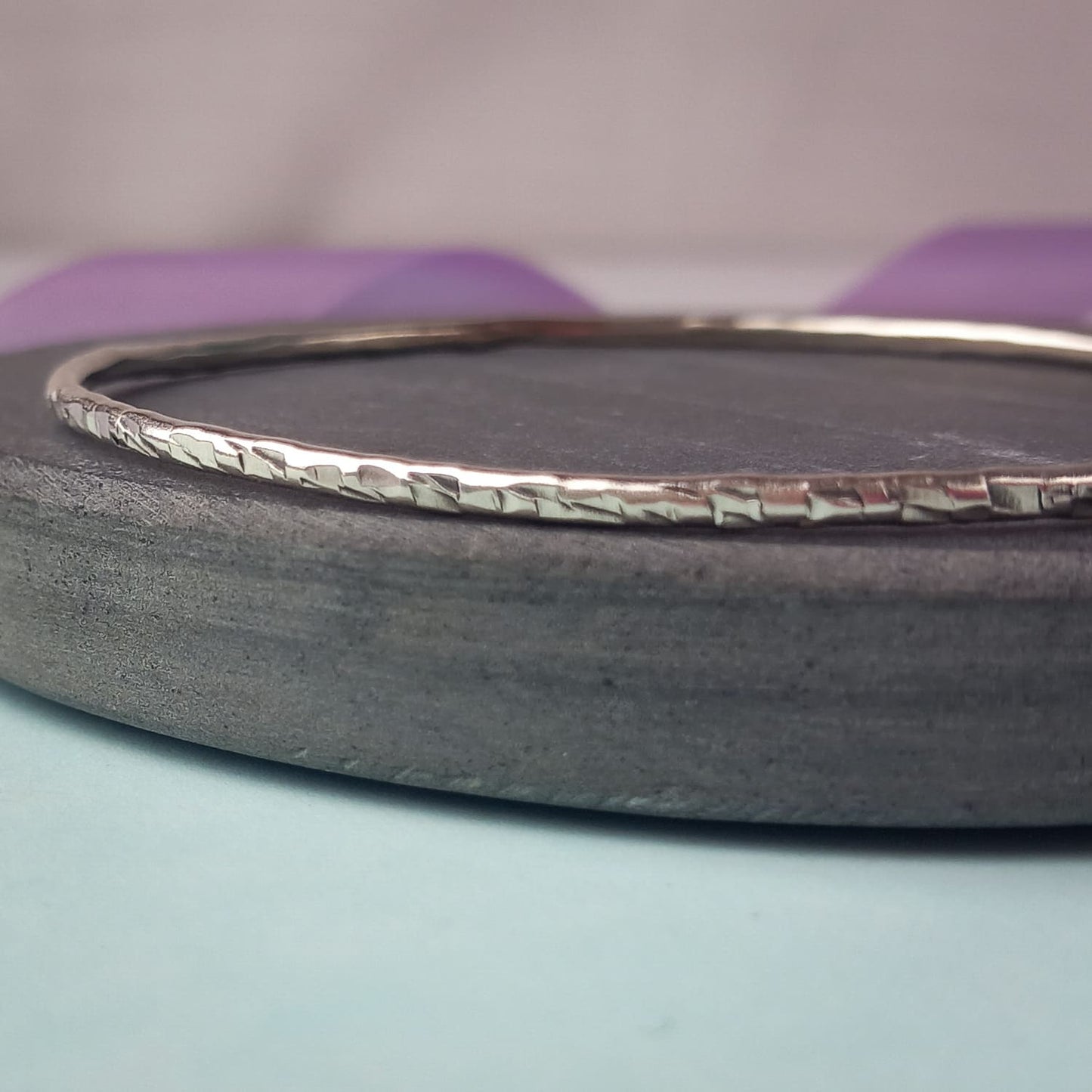 Image shows a heavily textured silver bangle sitting on a piece of dark grey slate. In the background is a lavender ribbon, adding a touch of colour.
