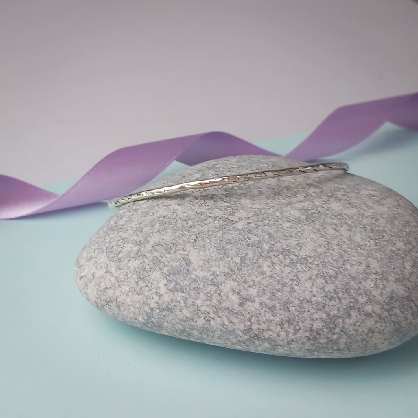 Image shows a heavily textured silver bangle sitting on a smooth, pale grey rock. In the background is a lavender ribbon, adding a touch of colour.