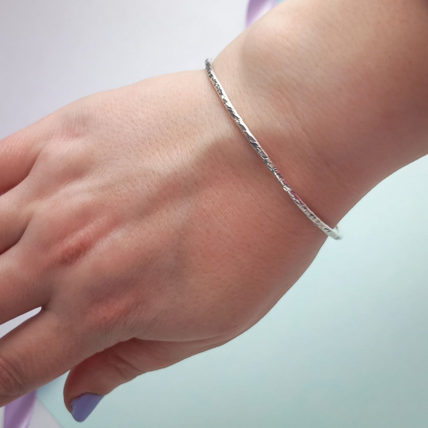 Image shows a heavily textured silver bangle being worn on a woman's wrist. The background is a soft, pale blue colour and she wears lavender nail polish, adding a touch of colour.