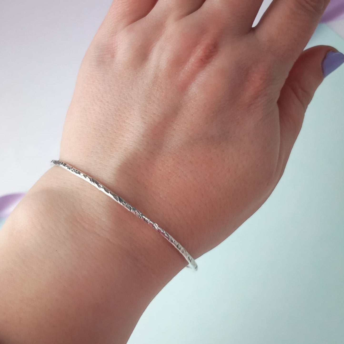 Image shows a heavily textured silver bangle being worn on a woman's wrist. The background is a soft, pale blue colour and she wears lavender nail polish, adding a touch of colour.