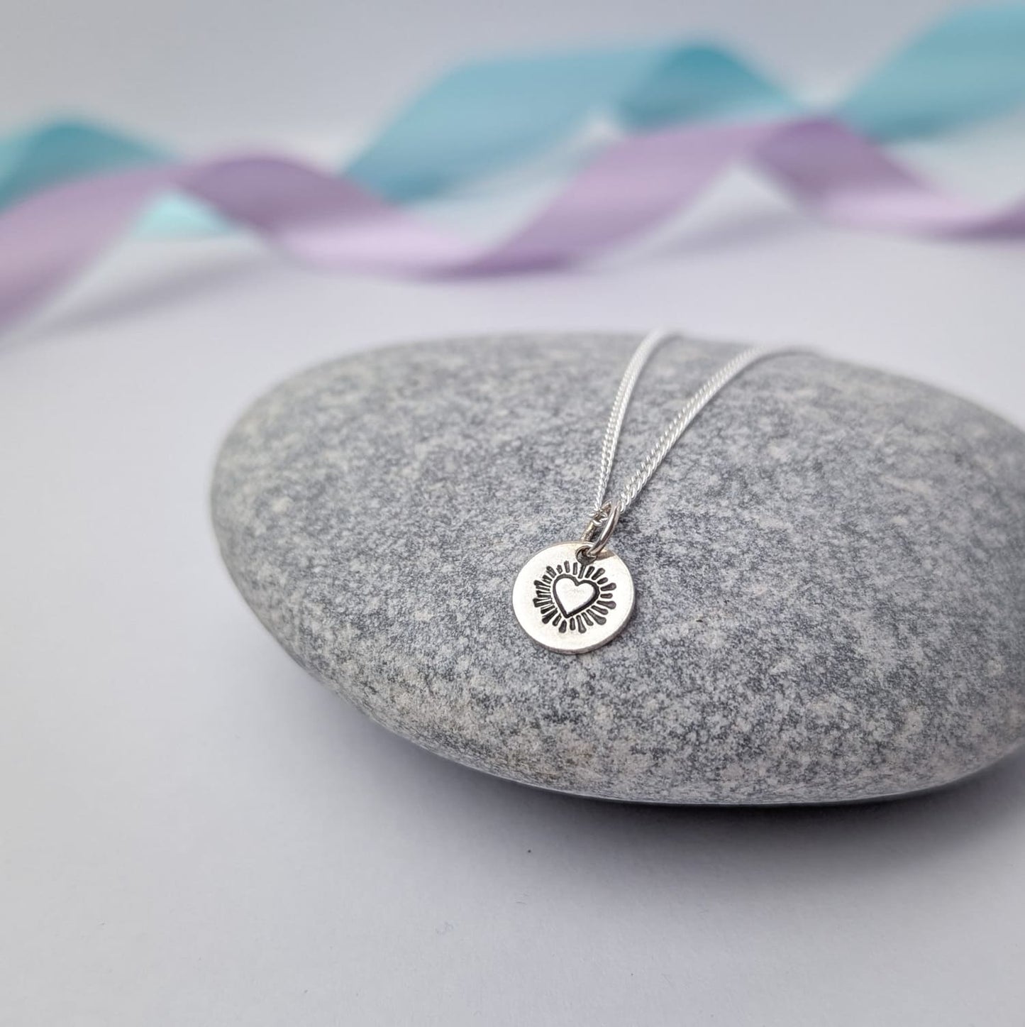 Photograph of a delicate silver pendant necklace resting on a smooth grey stone. The round disc pendant features a hand-stamped heart in the centre, surrounded by radiating lines resembling sun rays. The necklace has a fine silver chain. The background is softly blurred, with pastel blue and lavender ribbons adding a touch of colour.