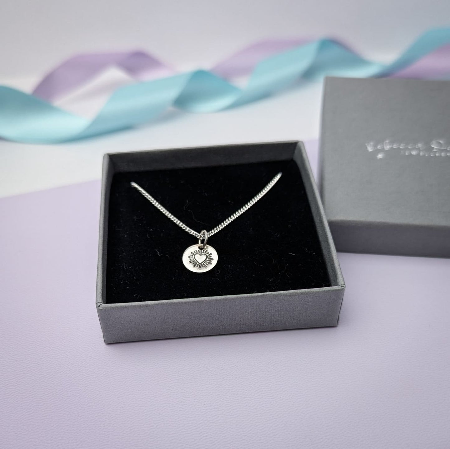 Photograph of a delicate silver pendant necklace in a charcoal and black presentation box. The round disc pendant features a hand-stamped heart in the centre, surrounded by radiating lines resembling sun rays. The necklace has a fine silver chain. The background is softly blurred, with pastel blue and lavender ribbons adding a touch of colour.