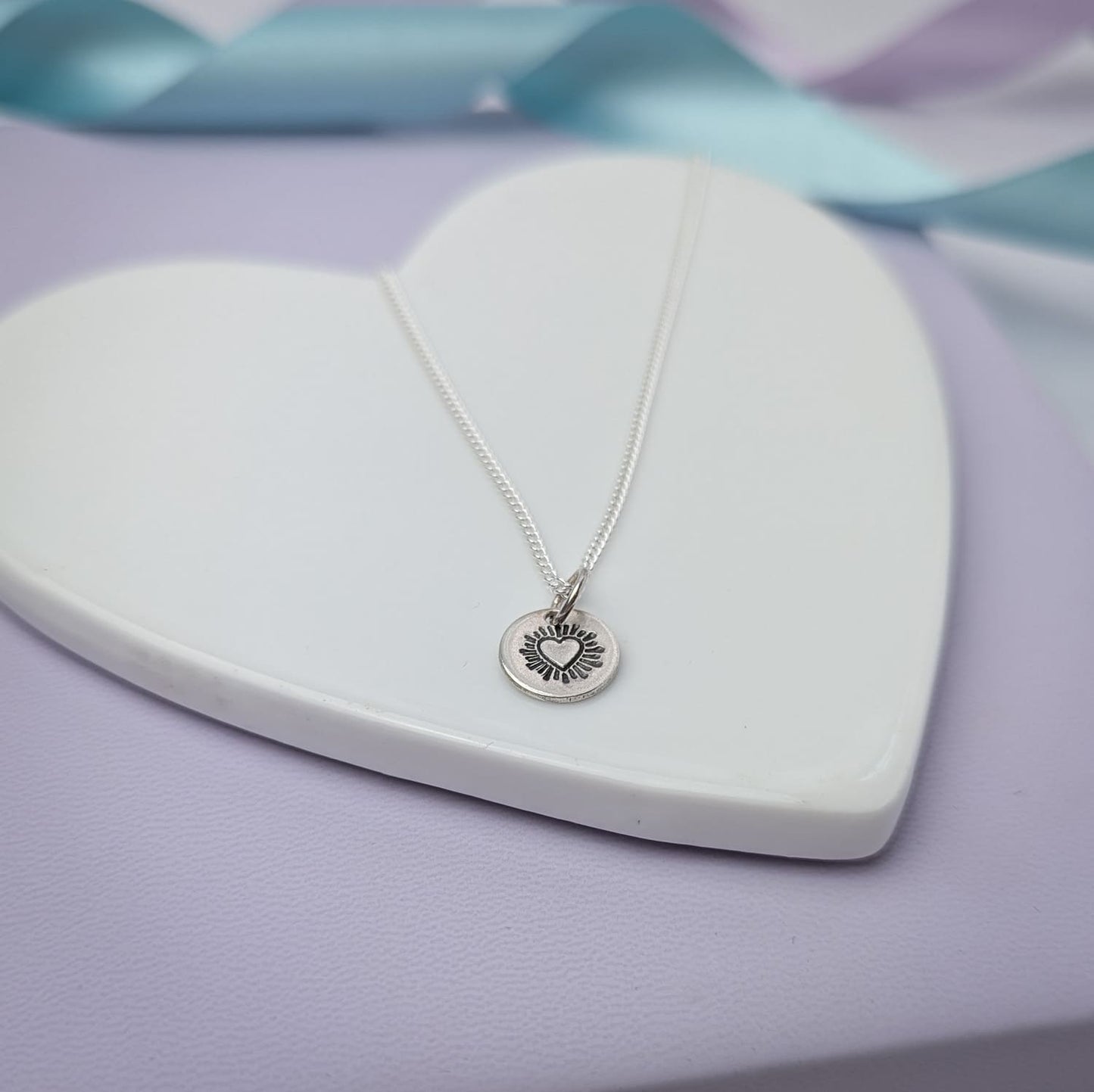 Photograph of a delicate silver pendant necklace resting on a white ceramic heart. The round disc pendant features a hand-stamped heart in the centre, surrounded by radiating lines resembling sun rays. The necklace has a fine silver chain. The background is softly blurred, with pastel blue and lavender ribbons adding a touch of colour.