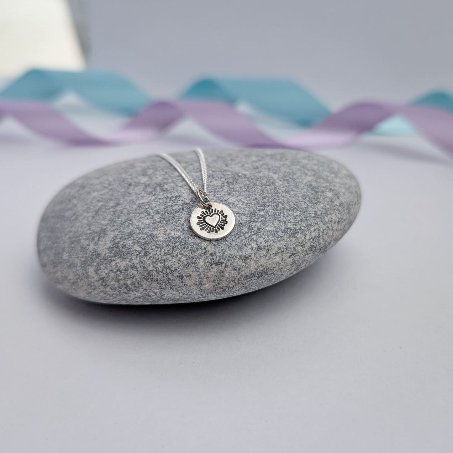 Photograph of a delicate silver pendant necklace resting on a smooth grey stone. The round disc pendant features a hand-stamped heart in the centre, surrounded by radiating lines resembling sun rays. The necklace has a fine silver chain. The background is softly blurred, with pastel blue and lavender ribbons adding a touch of colour.