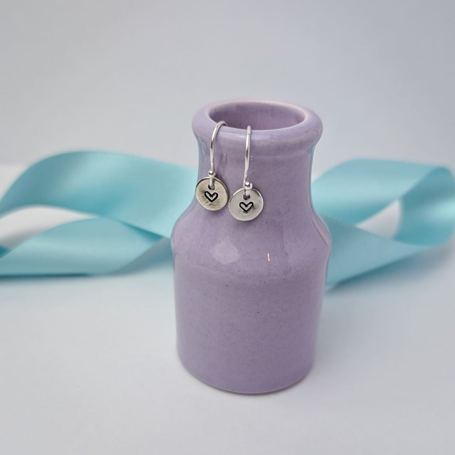 Photograph of a delicate pair of silver dangly earrings, resting on a tiny purple vase. Each earring features a round disc with a hand-stamped heart in the centre. The background is softly blurred, with pastel blue ribbons adding a touch of colour.