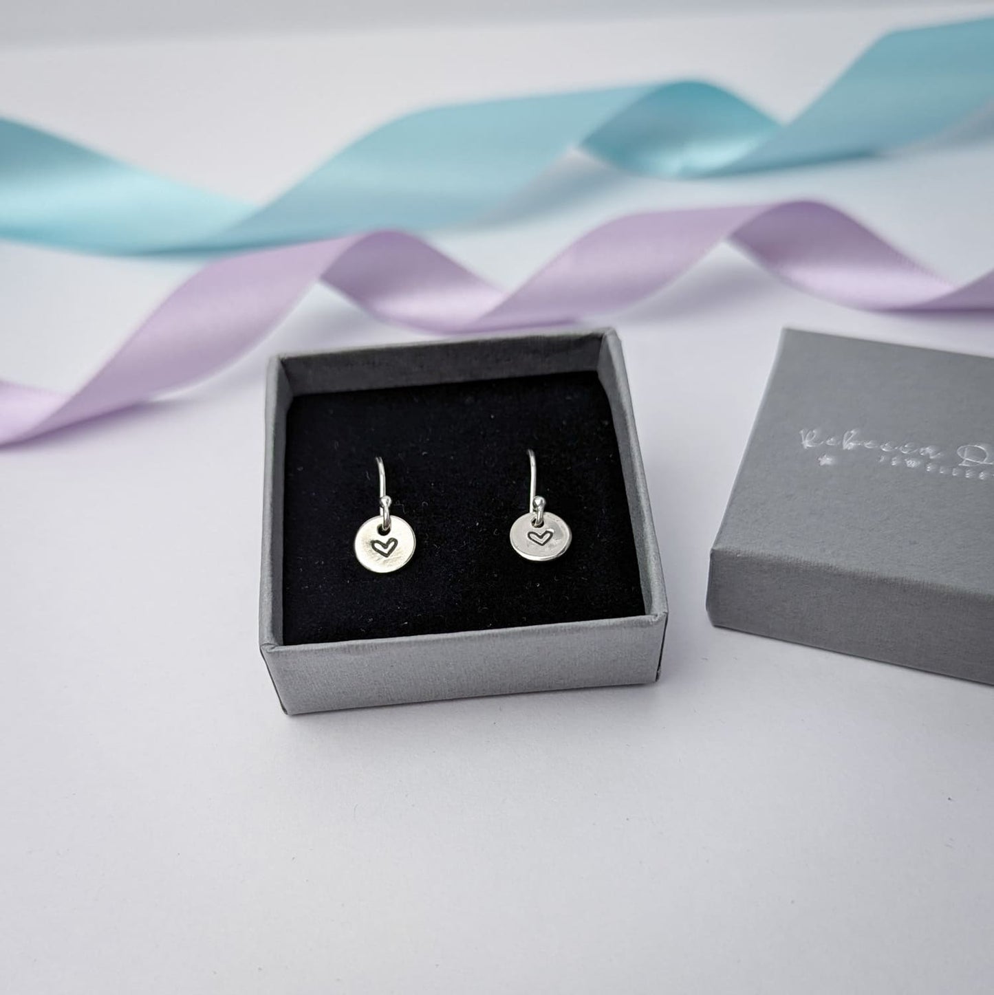 Photograph of a delicate pair of silver dangly earrings, displayed in a charcoal grey luxury gift box. Each earring features a round disc with a hand-stamped heart in the centre. The background is softly blurred, with pastel blue and lavender ribbons adding a touch of colour.