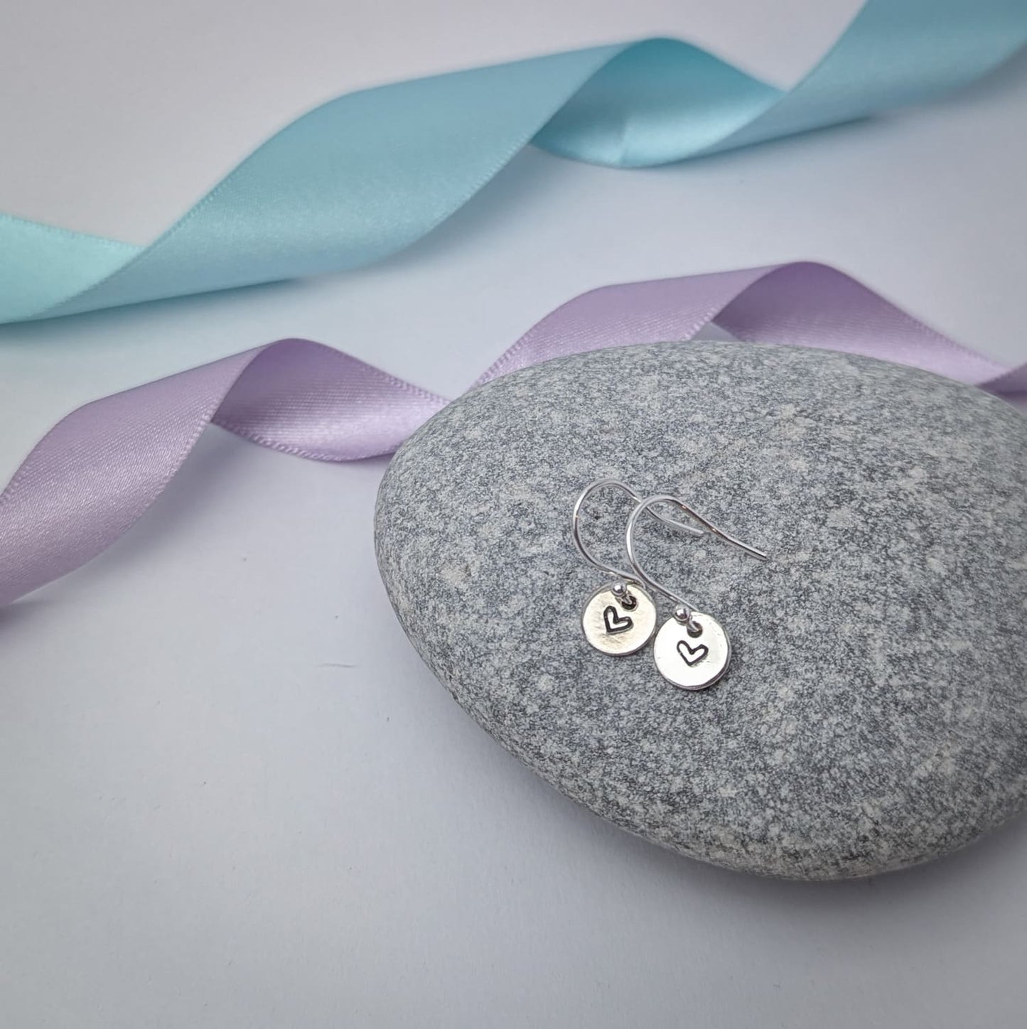 Photograph of a delicate pair of silver dangly earrings, resting on a smooth grey stone. Each earring features a round disc with a hand-stamped heart in the centre. The background is softly blurred, with pastel blue and lavender ribbons adding a touch of colour.