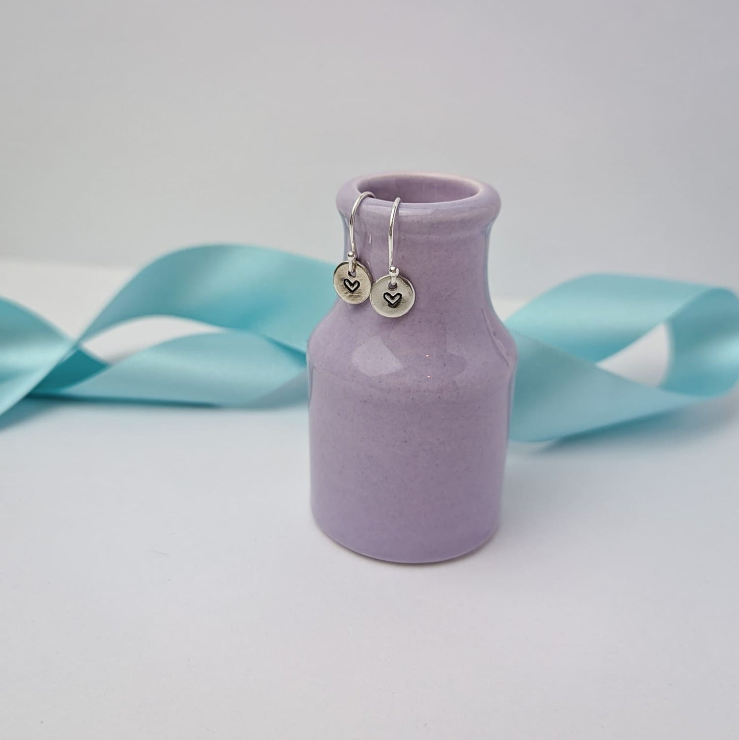 Photograph of a delicate pair of silver dangly earrings, resting on a tiny purple vase. Each earring features a round disc with a hand-stamped heart in the centre. The background is softly blurred, with pastel blue ribbons adding a touch of colour.