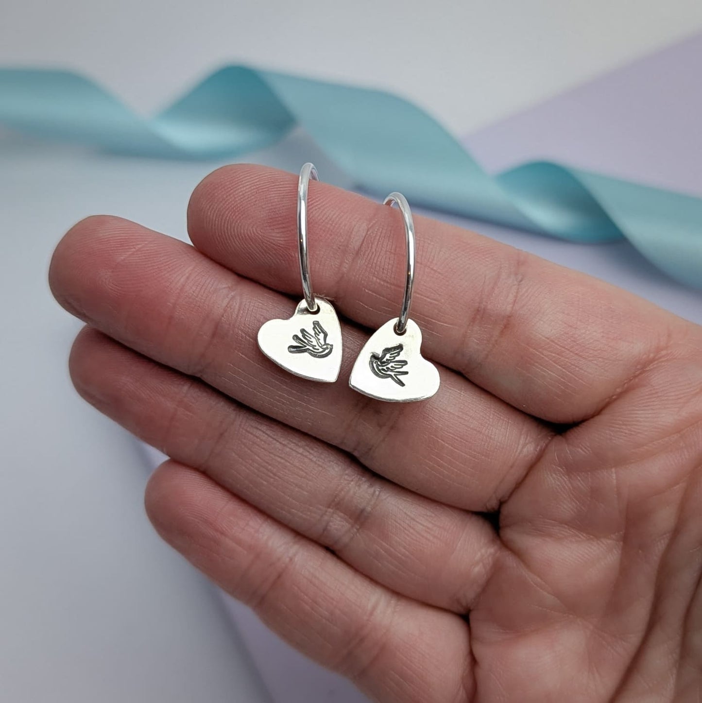 Photograph of a delicate pair of silver hoop earrings, held in a woman's hand. Each earring features a heart shaped charm with a hand-stamped tattoo-style swallow in the centre. The birds face each other in a mirror image. The background is softly blurred, with pastel blue ribbons adding a touch of colour.