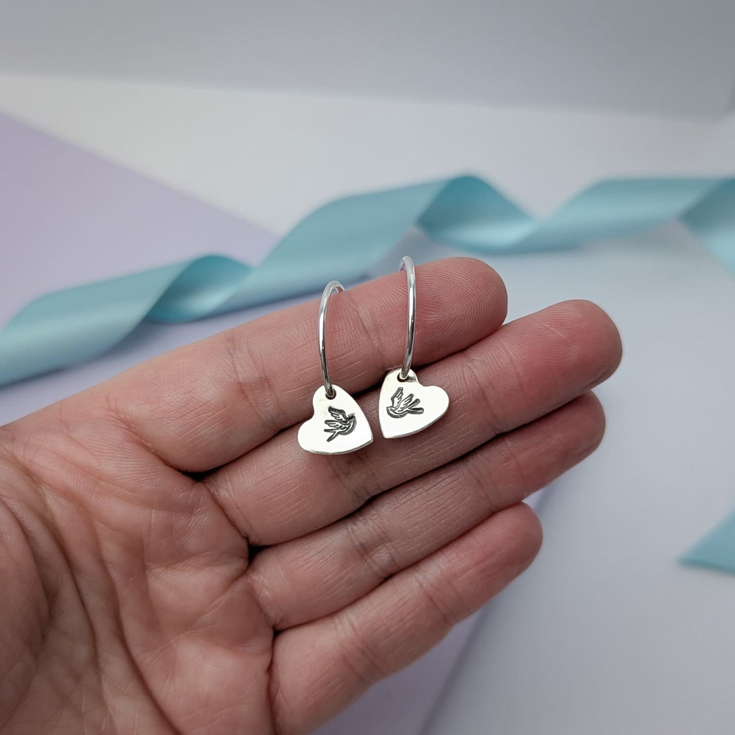 Photograph of a delicate pair of silver hoop earrings, held in a woman's hand. Each earring features a heart shaped charm with a hand-stamped tattoo-style swallow in the centre. The birds face each other in a mirror image. The background is softly blurred, with pastel blue ribbons adding a touch of colour.