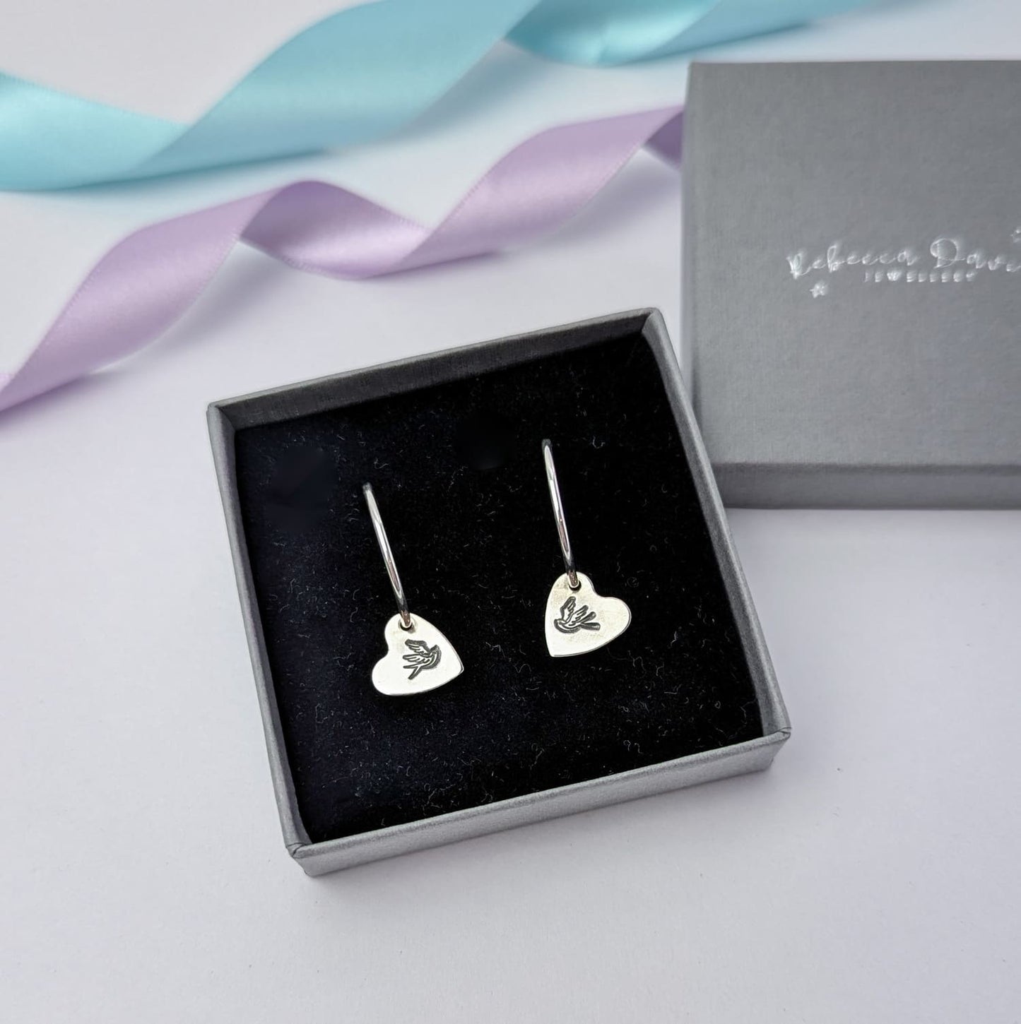 Photograph of a delicate pair of silver hoop earrings, displayed in a luxury charcoal grey gift box. Each earring features a heart shaped charm with a hand-stamped tattoo-style swallow in the centre. The birds face each other in a mirror image. The background is softly blurred, with pastel blue and lavender ribbons adding a touch of colour.