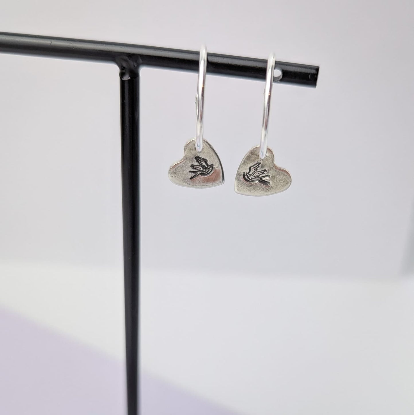 Close up photograph of a delicate pair of silver hoop earrings, displayed on a black earring holder. Each earring features a heart shaped charm with a hand-stamped tattoo-style swallow in the centre. The birds face each other in a mirror image.