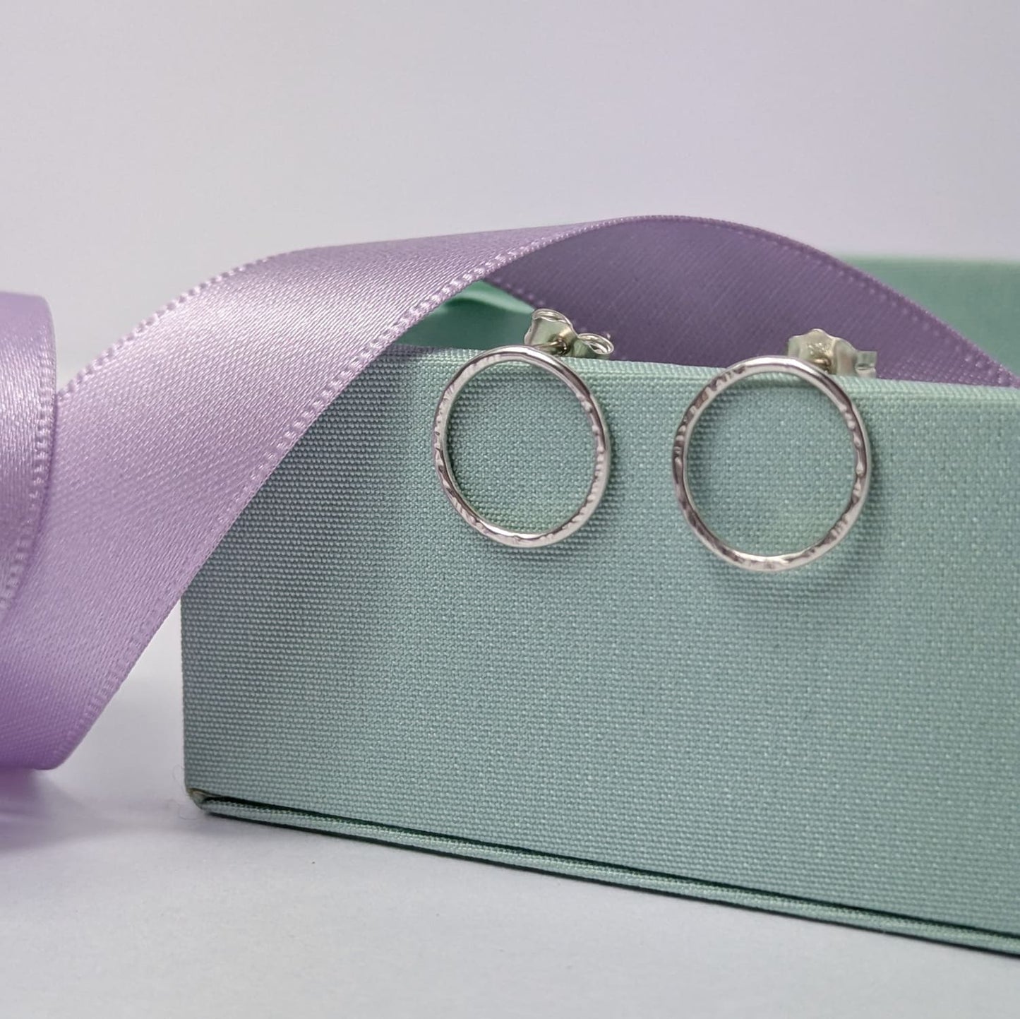 Photograph of a pair of silver circle stud earrings resting on a pastel green block. They have a hammered finish and an open design. The background is softly blurred, with pastel lavender ribbons adding a touch of colour.