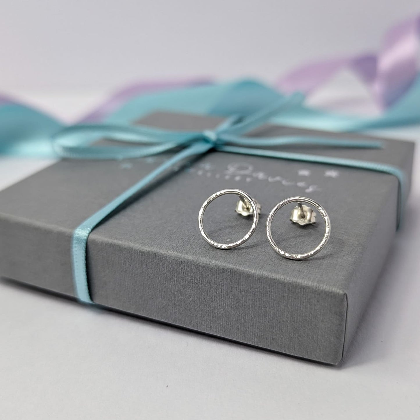 Photograph of a pair of silver circle stud earrings resting on top of a luxury charcoal grey gift box. They have a hammered finish and an open design. The background is softly blurred, with pastel blue and lavender ribbons adding a touch of colour.