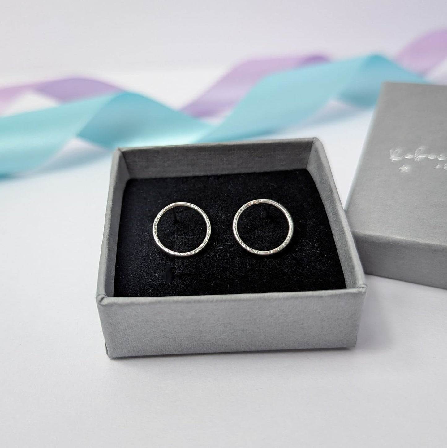 Photograph of a pair of silver circle stud earrings displayed in a grey gift box. They have a hammered finish and an open design. The background is softly blurred, with pastel lavender ribbons adding a touch of colour.