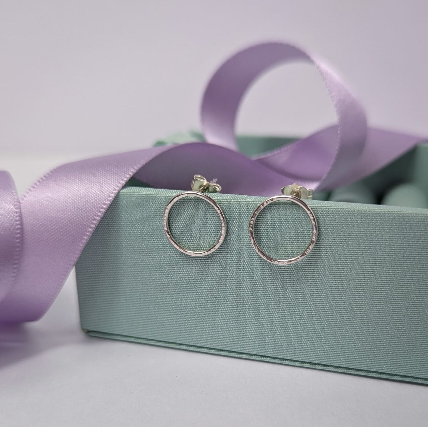Photograph of a pair of silver circle stud earrings resting on a pastel green block. They have a hammered finish and an open design. The background is softly blurred, with pastel lavender ribbons adding a touch of colour.
