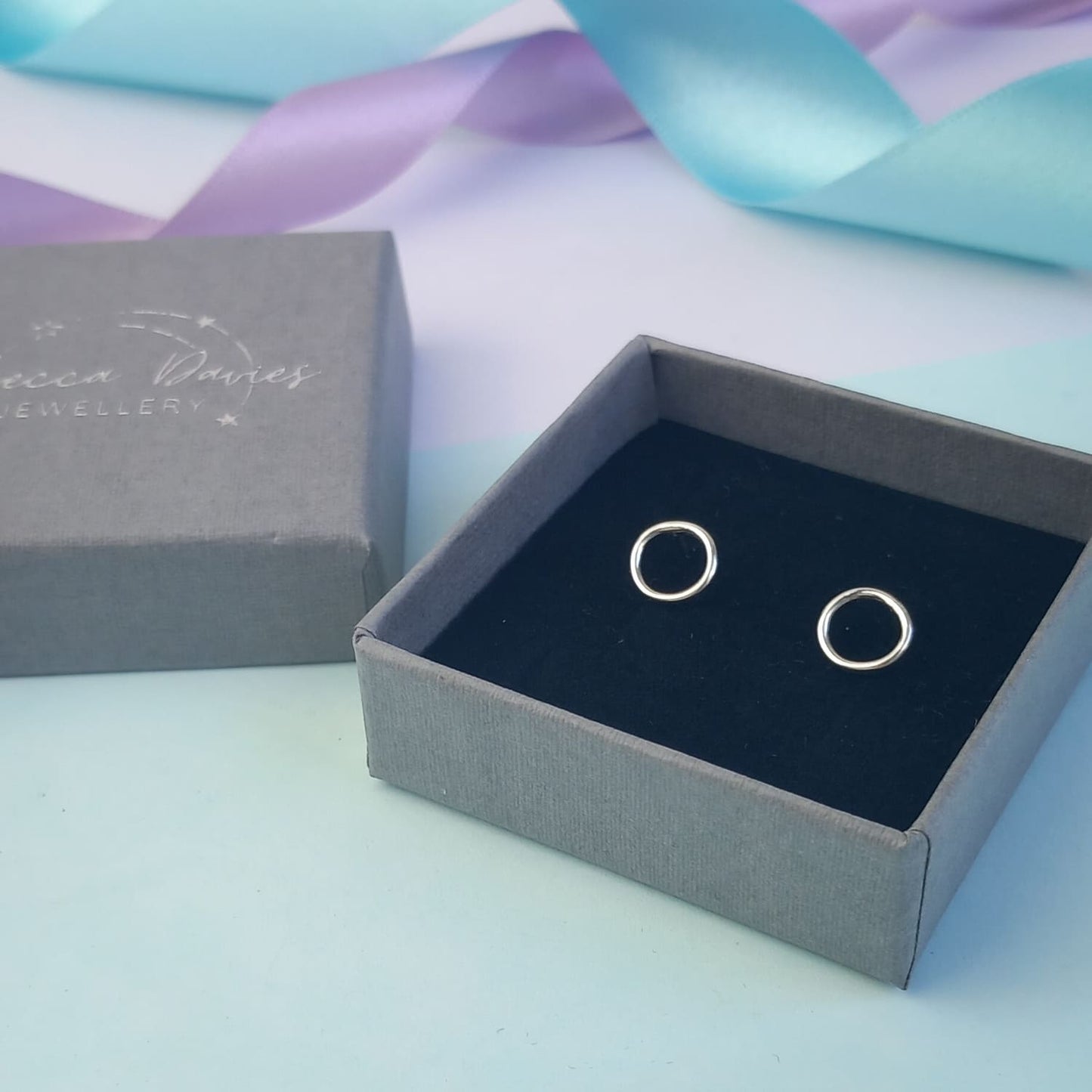 Photograph of a pair of silver circle stud earrings displayed in a luxury charcoal grey gift box. They have a polished finish and an open design. The background is softly blurred, with pastel lavender ribbons adding a touch of colour.