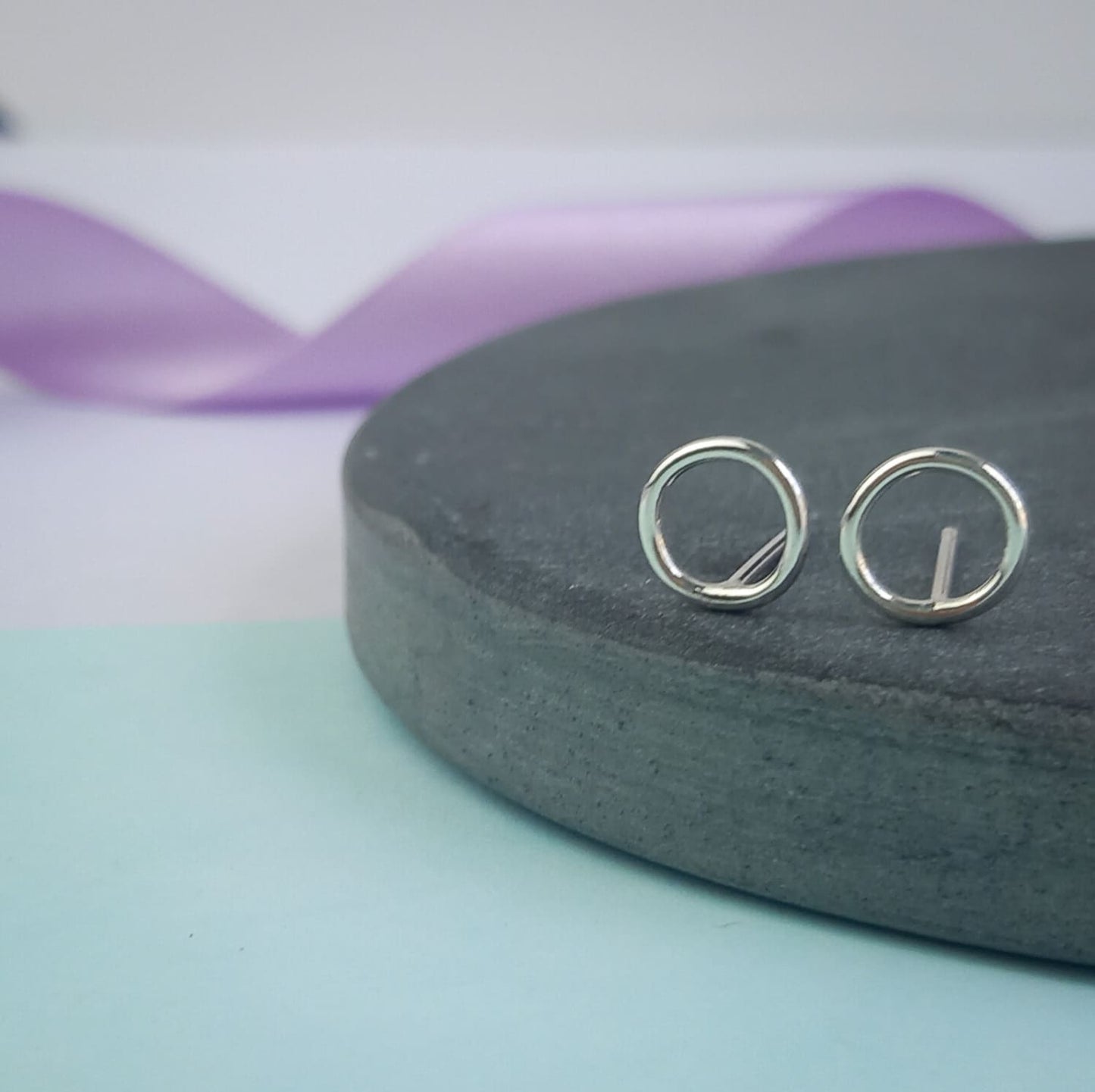 Photograph of a pair of silver circle stud earrings resting on a slate disc. They have a polished finish and an open design. The background is softly blurred, with pastel lavender ribbons adding a touch of colour.
