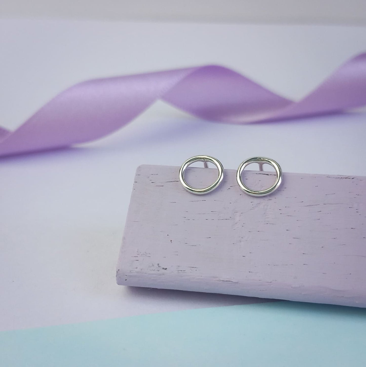 Photograph of a pair of silver circle stud earrings resting on a purple block. They have a polished finish and an open design. The background is softly blurred, with pastel lavender ribbons adding a touch of colour.
