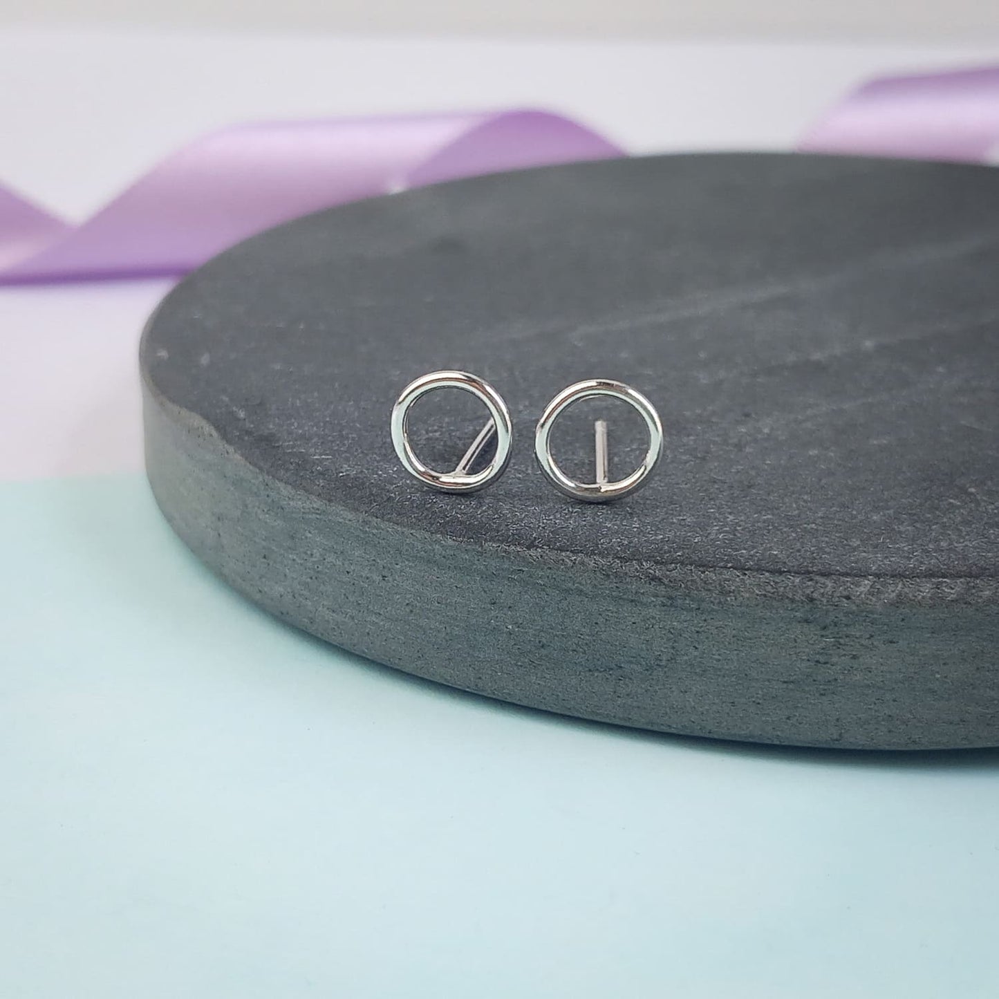 Photograph of a pair of silver circle stud earrings resting on a slate disc. They have a polished finish and an open design. The background is softly blurred, with pastel lavender ribbons adding a touch of colour.