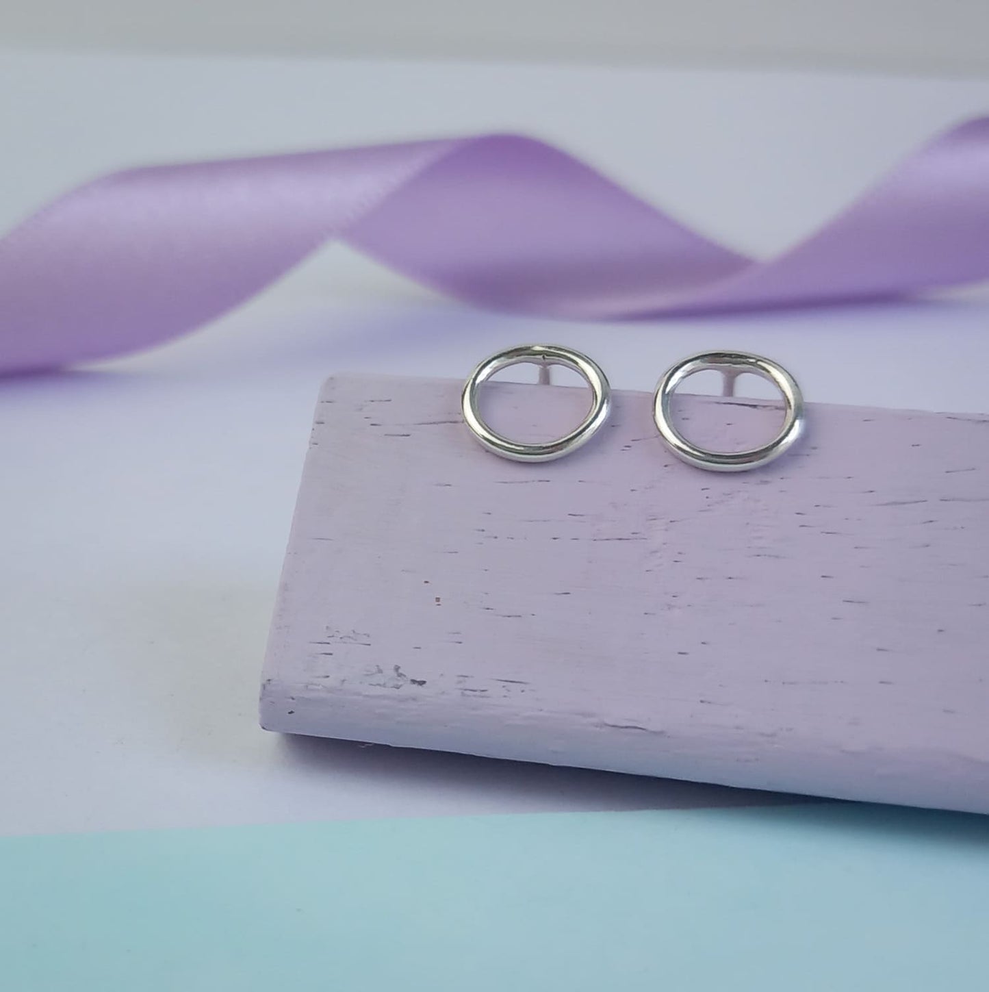 Photograph of a pair of silver circle stud earrings resting on a purple block. They have a polished finish and an open design. The background is softly blurred, with pastel lavender ribbons adding a touch of colour.