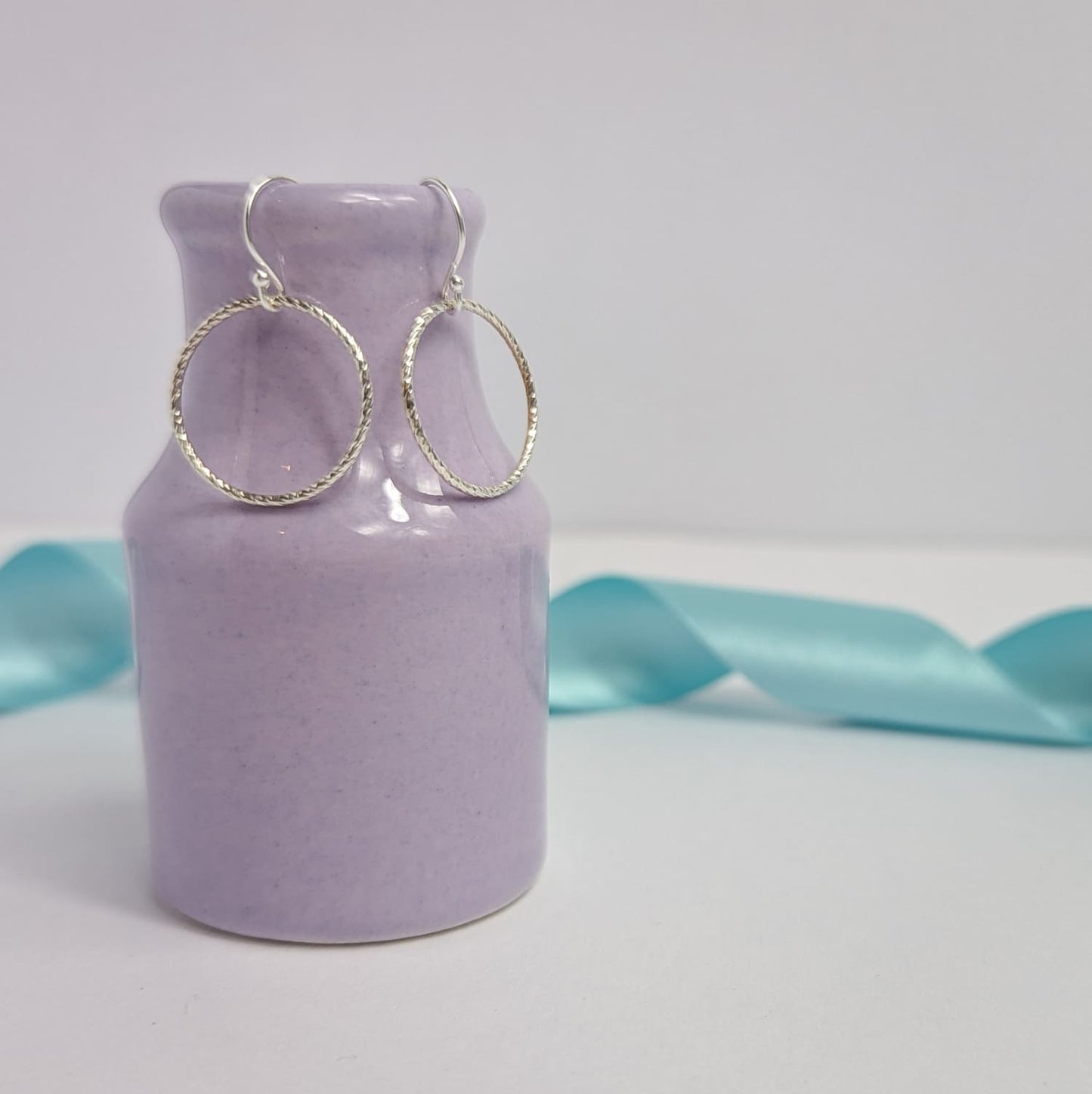 Photograph of a pair of silver open circle earrings hanging from the edge of a tiny purple vase. Each earring features an open circle with a soft hammered texture that beautifully catches the light. The background is softly blurred, with pastel blue ribbons adding a touch of colour.
