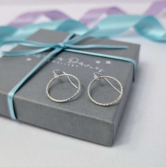 Photograph of a pair of silver open circle earrings resting on top of a closed luxury gift box tied with a turquoise ribbon. Each earring features an open circle with a soft hammered texture that beautifully catches the light. The background is softly blurred, with pastel blue and lavender ribbons adding a touch of colour.