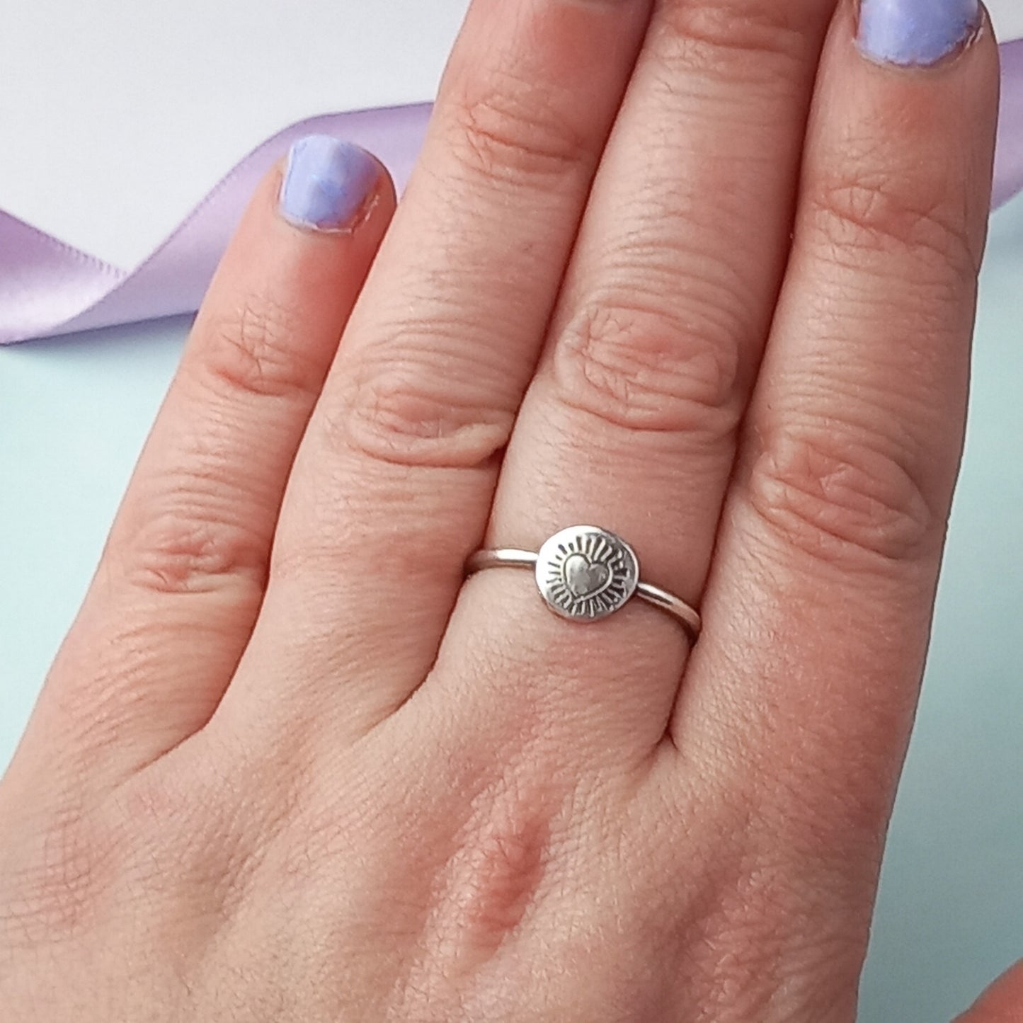 Photograph of a delicate silver ring being worn on the middle finger of a woman's hand. The ring features a round disc with a hand-stamped heart in the centre, surrounded by radiating lines resembling sun rays. The background is softly blurred, with lavender ribbons and the woman's nails are painted a lavender colour adding a touch of colour.