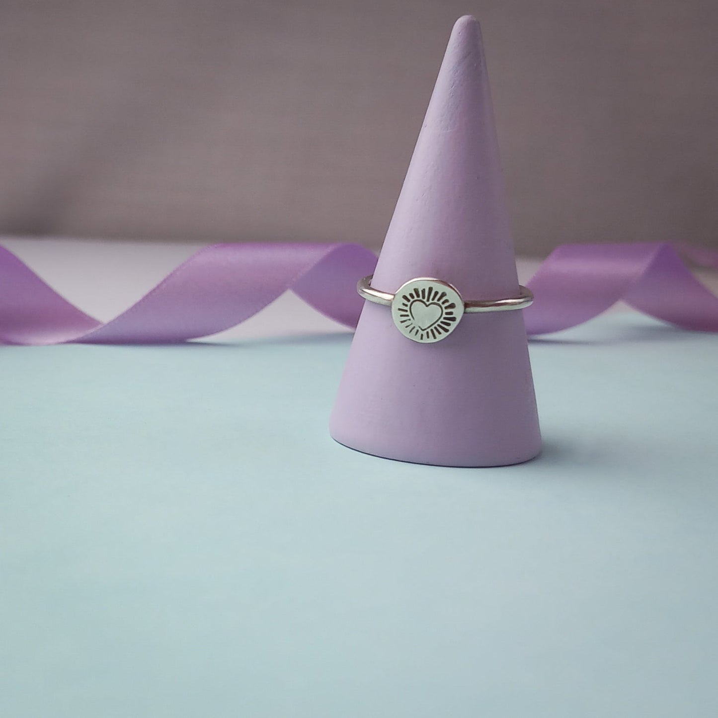 Photograph of a delicate silver ring resting on a soft lilac coloured ring holder. The ring features a round disc with a hand-stamped heart in the centre, surrounded by radiating lines resembling sun rays. The background is softly blurred, with pastel blue and lavender ribbons adding a touch of colour.
