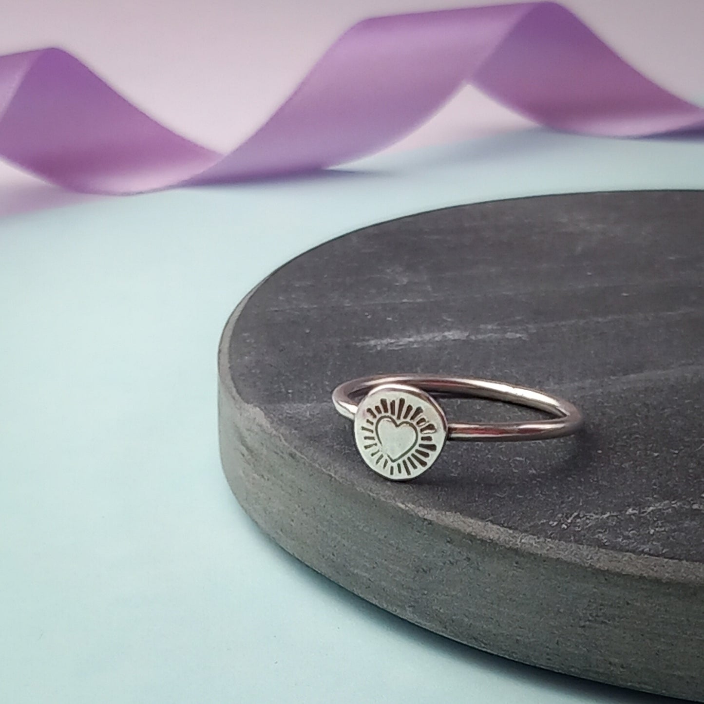 Photograph of a delicate silver ring resting on a dark slate. The ring features a round disc with a hand-stamped heart in the centre, surrounded by radiating lines resembling sun rays. The background is softly blurred, with pastel blue and lavender ribbons adding a touch of colour.