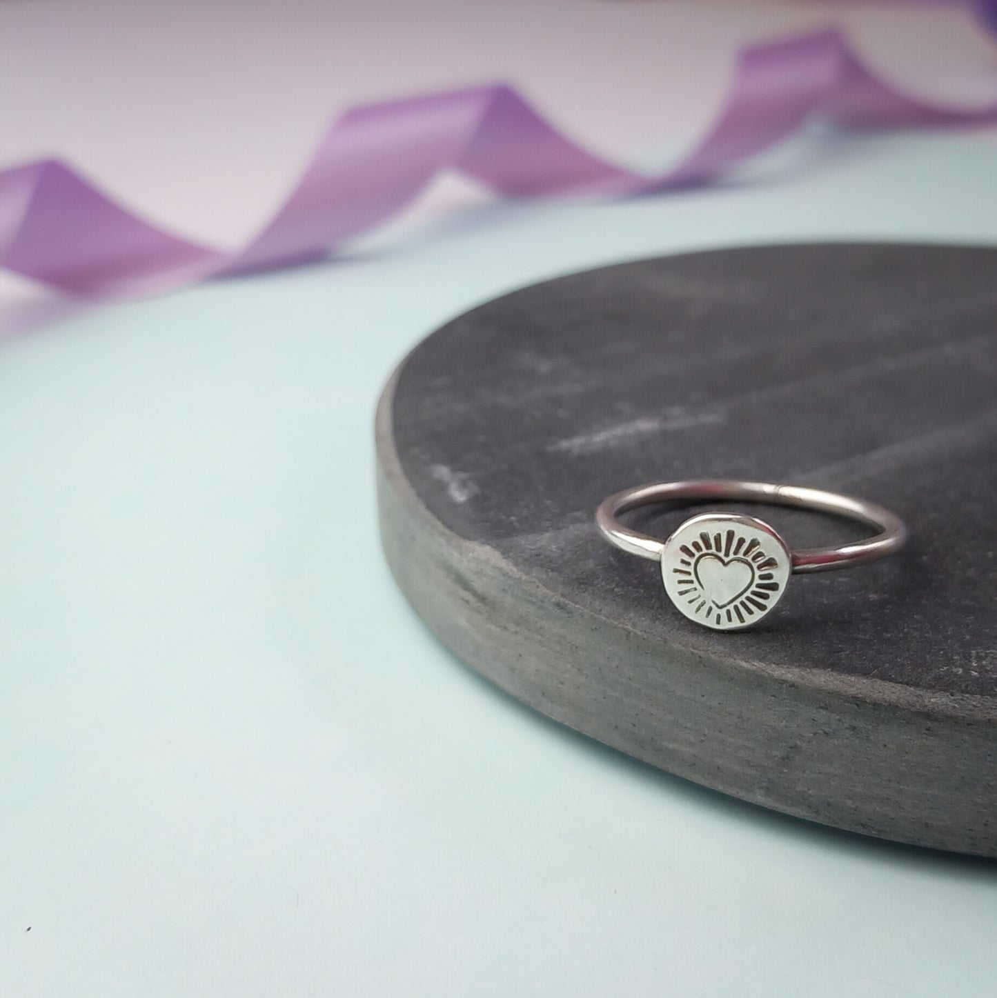 Photograph of a delicate silver ring resting on a dark slate. The ring features a round disc with a hand-stamped heart in the centre, surrounded by radiating lines resembling sun rays. The background is softly blurred, with pastel blue and lavender ribbons adding a touch of colour.