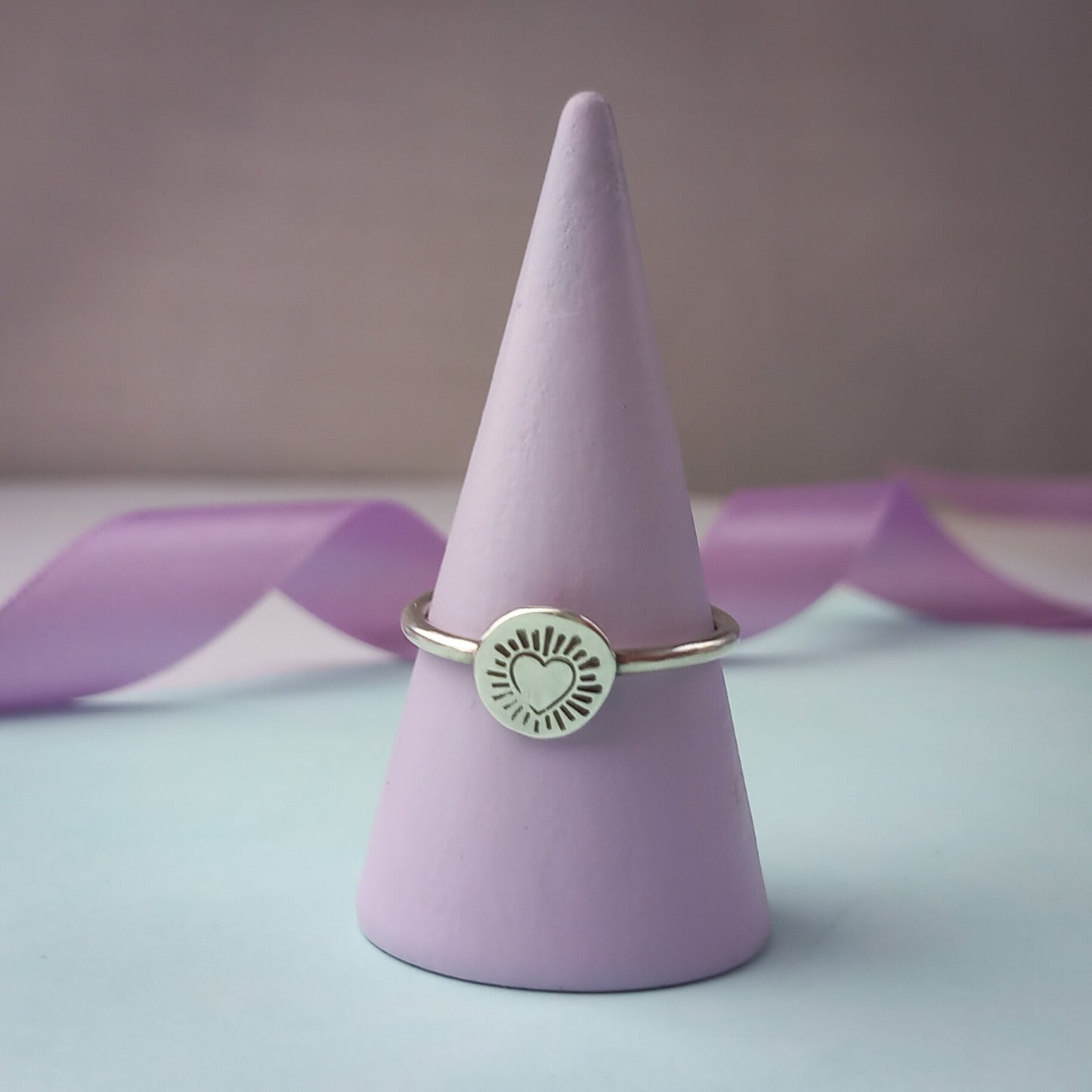 Photograph of a delicate silver ring resting on a soft lilac coloured ring holder. The ring features a round disc with a hand-stamped heart in the centre, surrounded by radiating lines resembling sun rays. The background is softly blurred, with pastel blue and lavender ribbons adding a touch of colour.