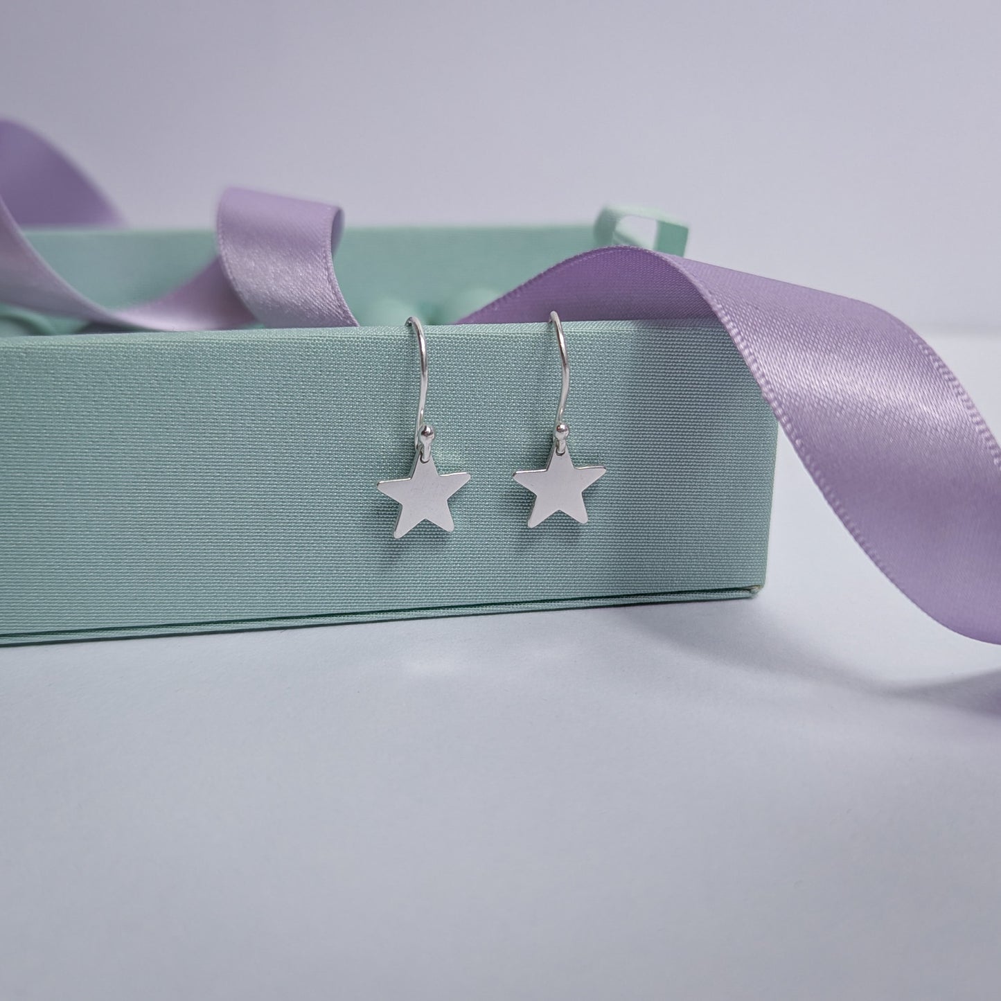 Handmade recycled silver star earrings displayed on a pastel box beside a soft purple ribbon, highlighting their shine and delicate craftsmanship.