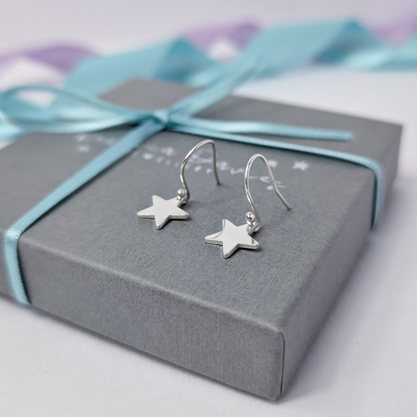 A pair of sterling silver star-shaped dangle earrings sit on top of a grey jewelry box, tied with a pastel ribbon. The box lid is embossed with the logo 'Rebecca Davies Jewellery'. The display is styled with pastel lavender and blue ribbons in the background.