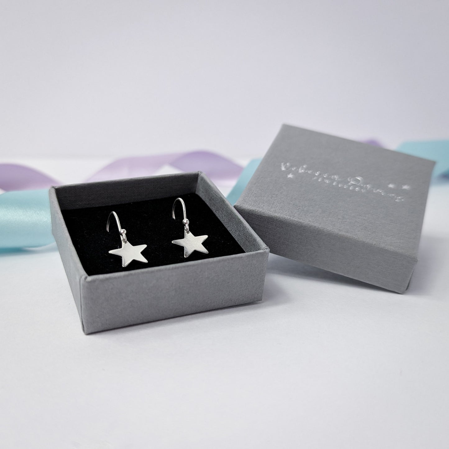 A pair of sterling silver star-shaped dangle earrings presented in a grey jewelry box with a black velvet interior. The box lid, embossed with the logo 'Rebecca Davies Jewellery,' rests slightly open. The display is styled with pastel lavender and blue ribbons in the background.