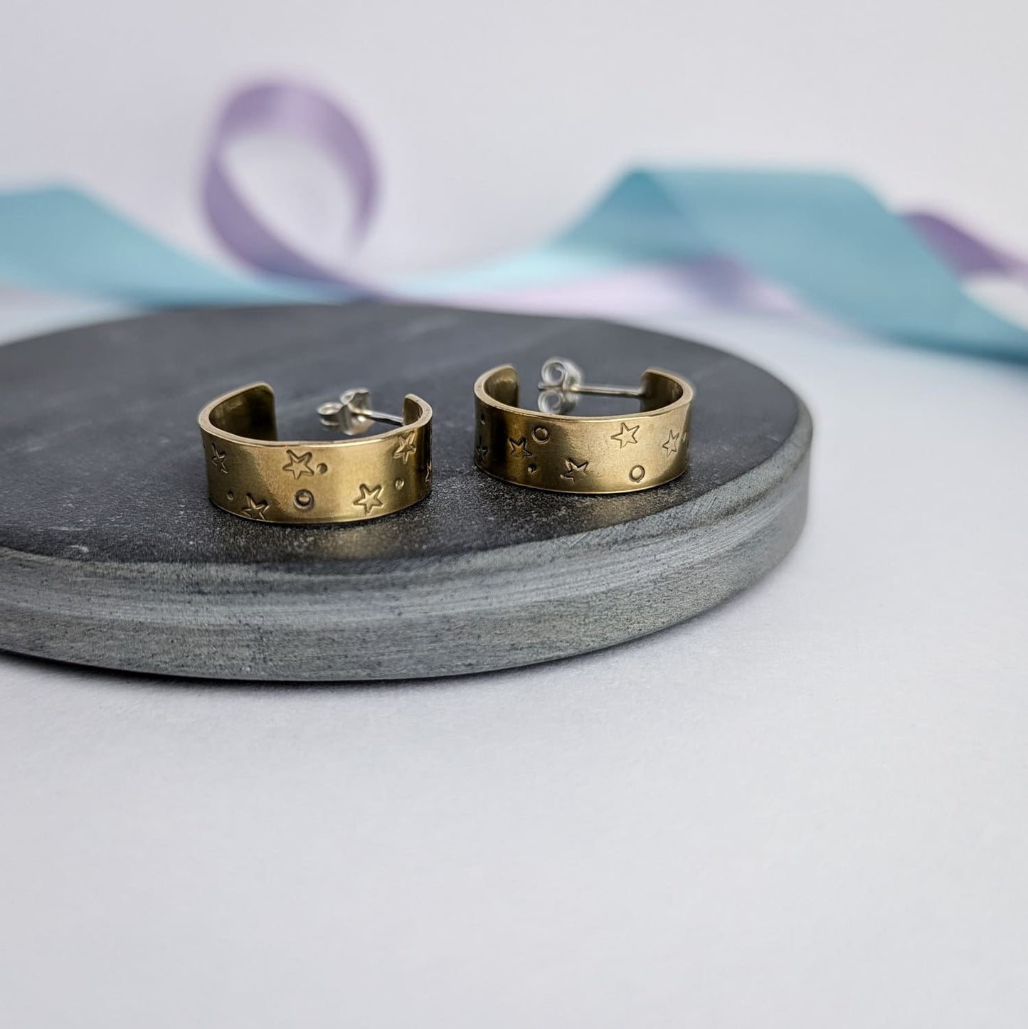 Image shows a pair of chunky brass hoop earrings with a stamped star and space design. The earrings are displayed on a dark slate circle. The background features pastel lavender and blue ribbons which add a touch of colour.