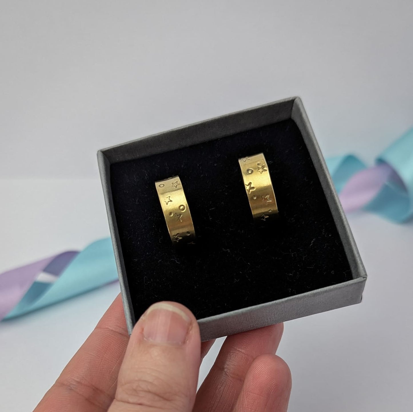 Image shows a pair of chunky brass hoop earrings with a stamped star and space design. The earrings are displayed in a dark grey gift box, held in a woman's hand. The background features pastel lavender and blue ribbons which add a touch of colour.