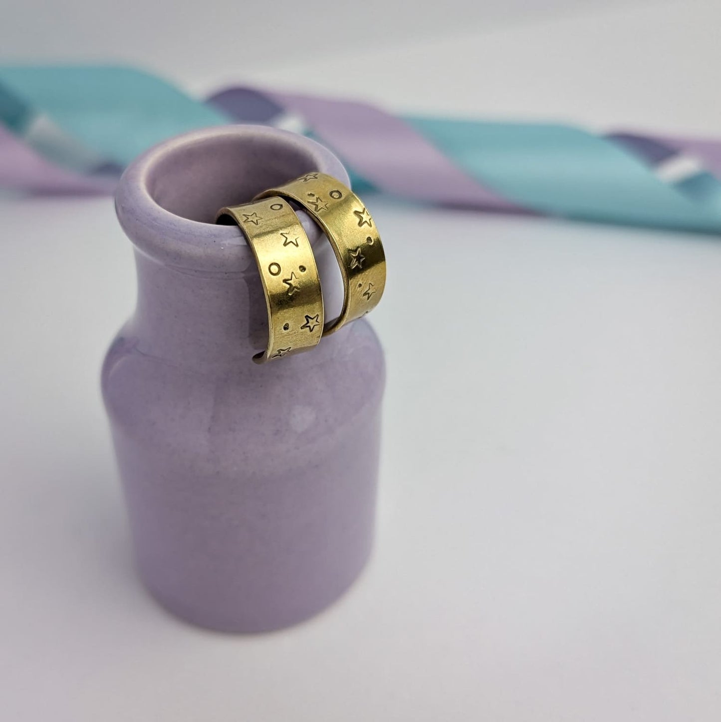 Image shows a pair of chunky brass hoop earrings with a stamped star and space design. The earrings are displayed hanging from the rim of a tiny purple vase. The background features pastel lavender and blue ribbons which add a touch of colour.