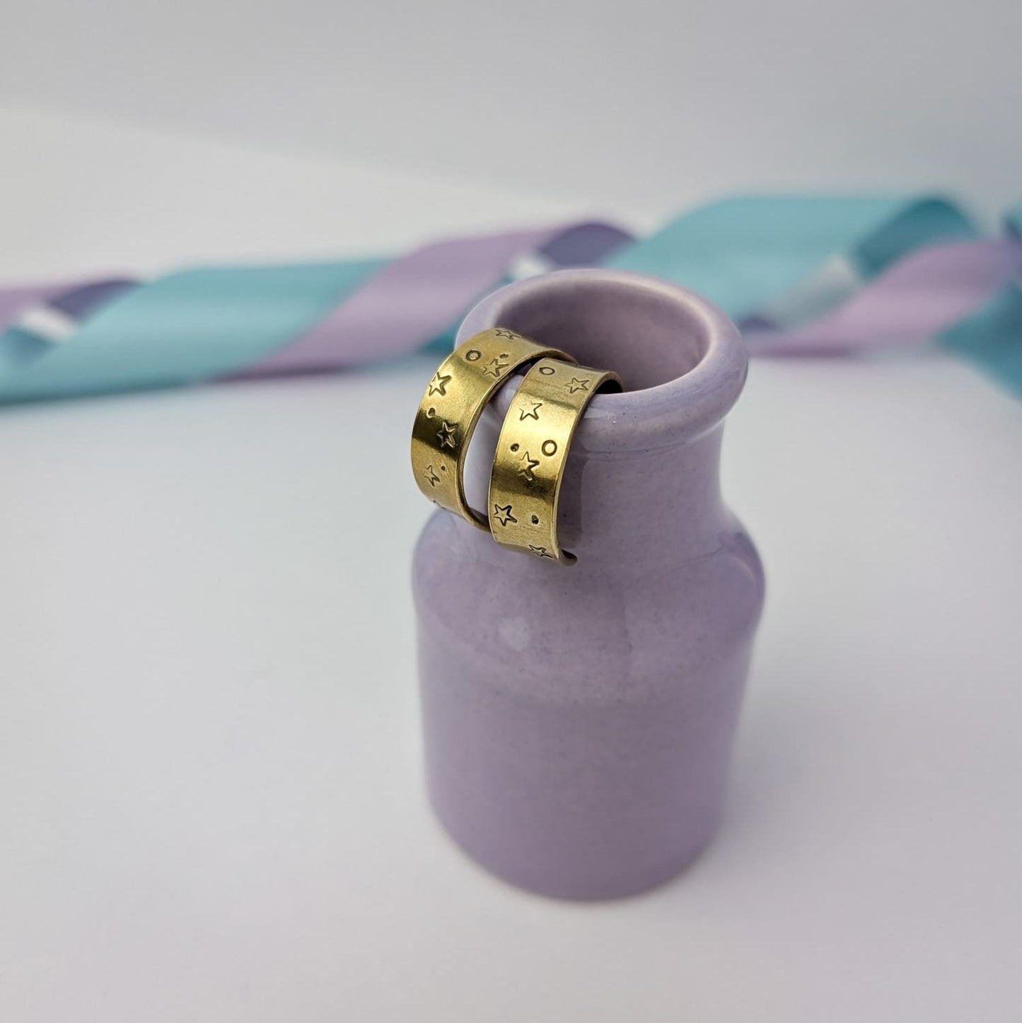 Image shows a pair of chunky brass hoop earrings with a stamped star and space design. The earrings are displayed hanging from the rim of a tiny purple vase. The background features pastel lavender and blue ribbons which add a touch of colour.