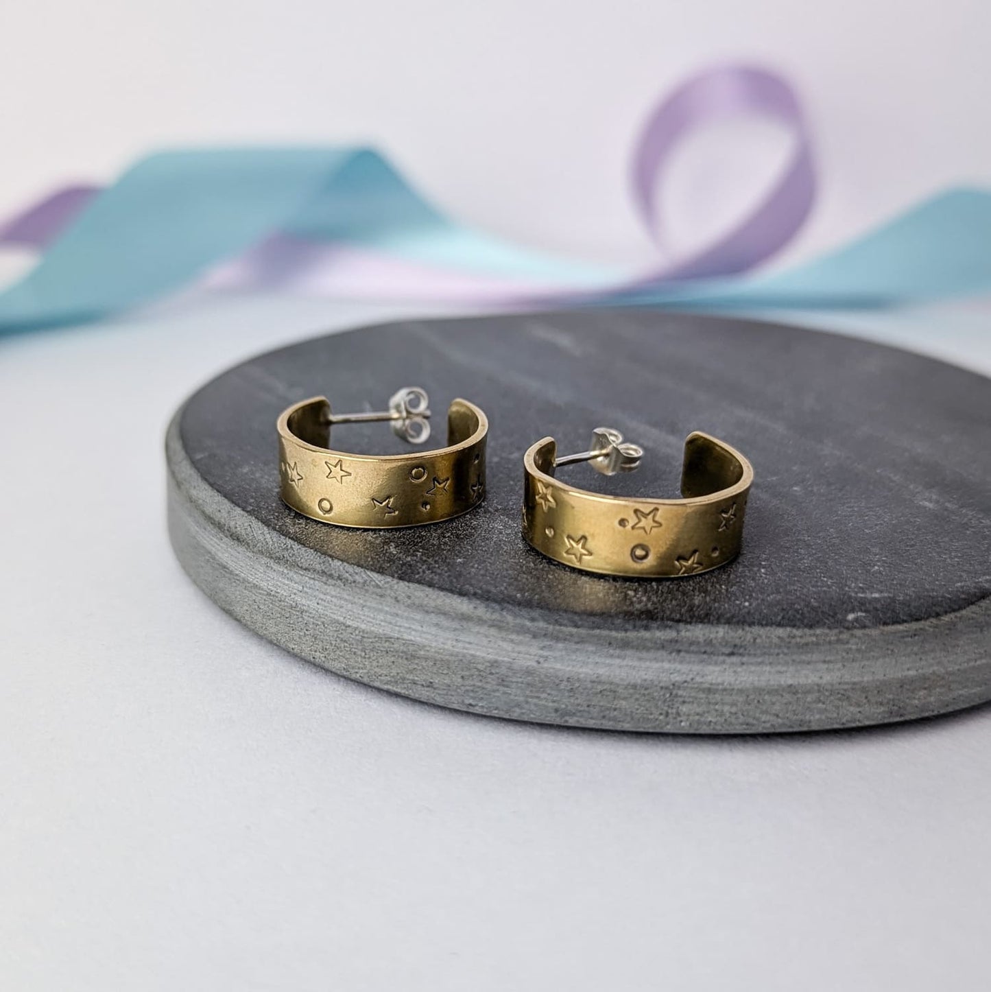 Image shows a pair of chunky brass hoop earrings with a stamped star and space design. The earrings are displayed on a dark slate circle. The background features pastel lavender and blue ribbons which add a touch of colour.