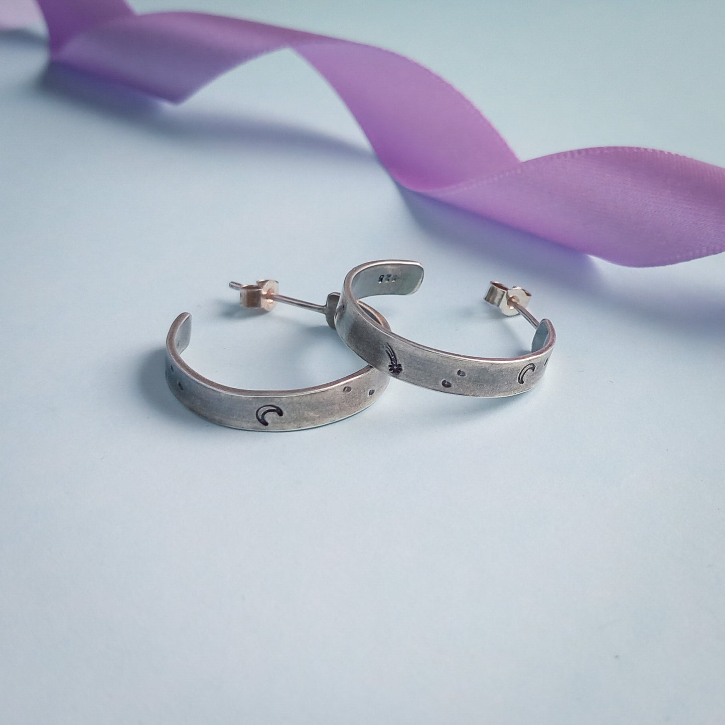 Two recycled silver chunky hoop earrings featuring stamped designs of a moon, comet and stars, elegantly displayed against a pastel background. Styled with a pastel lilac coloured ribbon in the background.