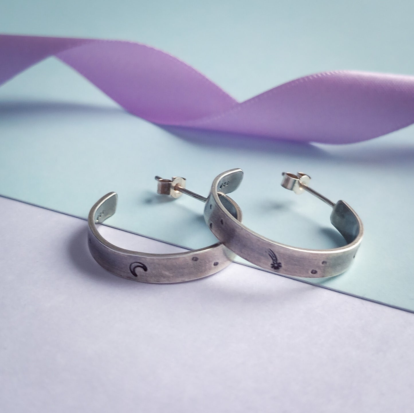 Two recycled silver chunky hoop earrings featuring stamped designs of a moon, comet and stars, elegantly displayed against a pastel background. Styled with a pastel lilac coloured ribbon in the background.